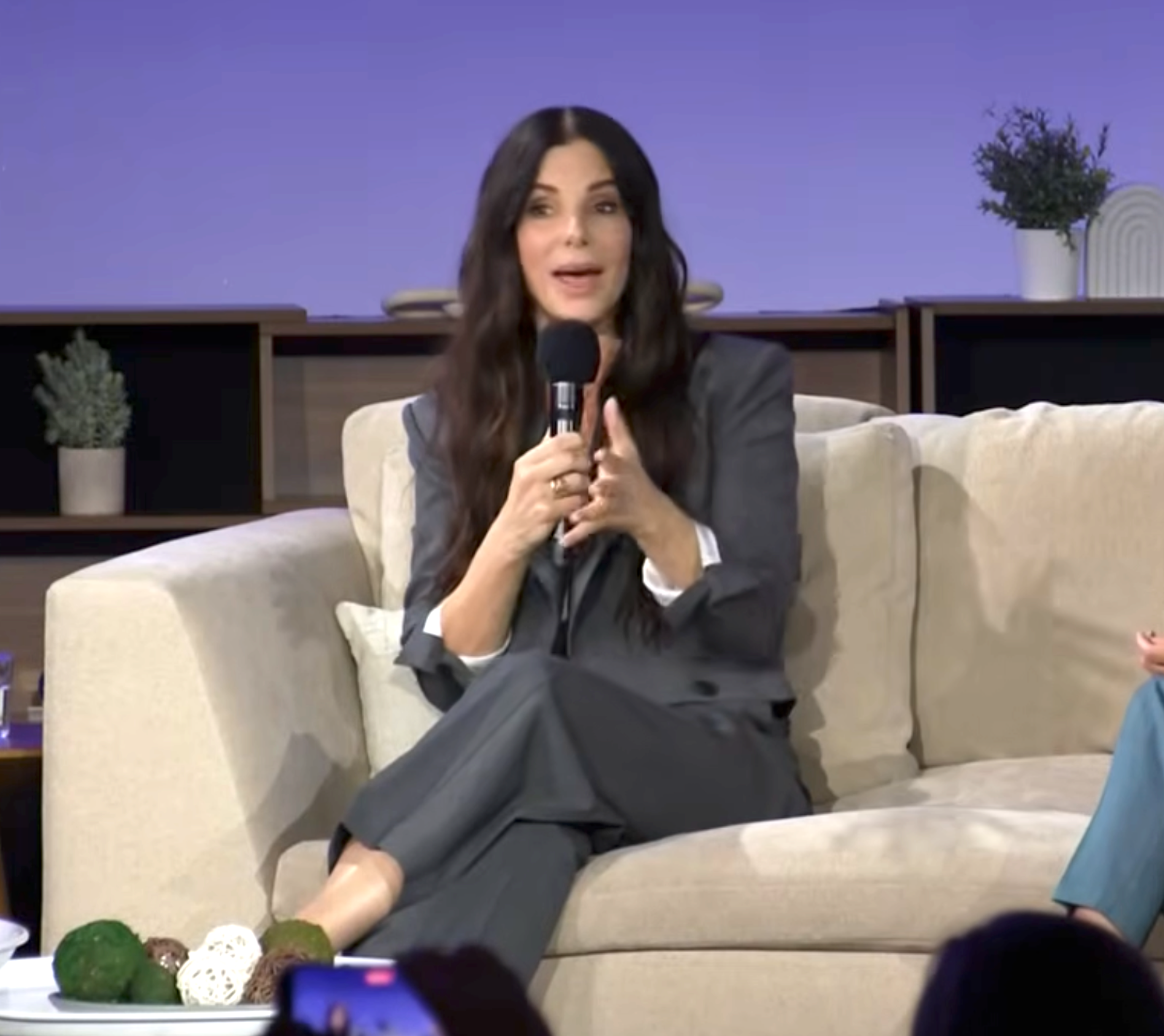 A idiosyncratic   successful  a tailored grey  suit   speaks into a microphone portion    seated connected  a couch, gesturing with 1  manus  during an event