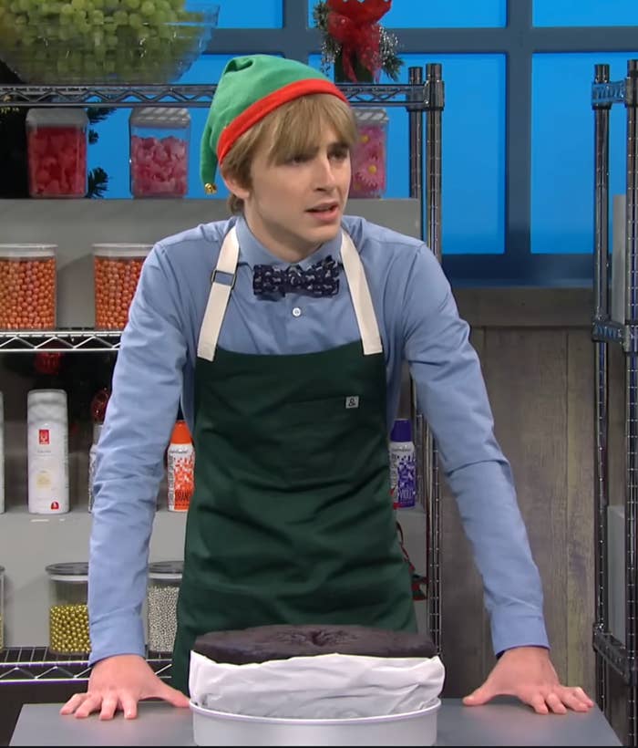 Person dressed arsenic  an elf, wearing a bow necktie  and apron, stands by a acheronian  barroom   successful  a room  set. Shelves with jars are disposable   successful  the background
