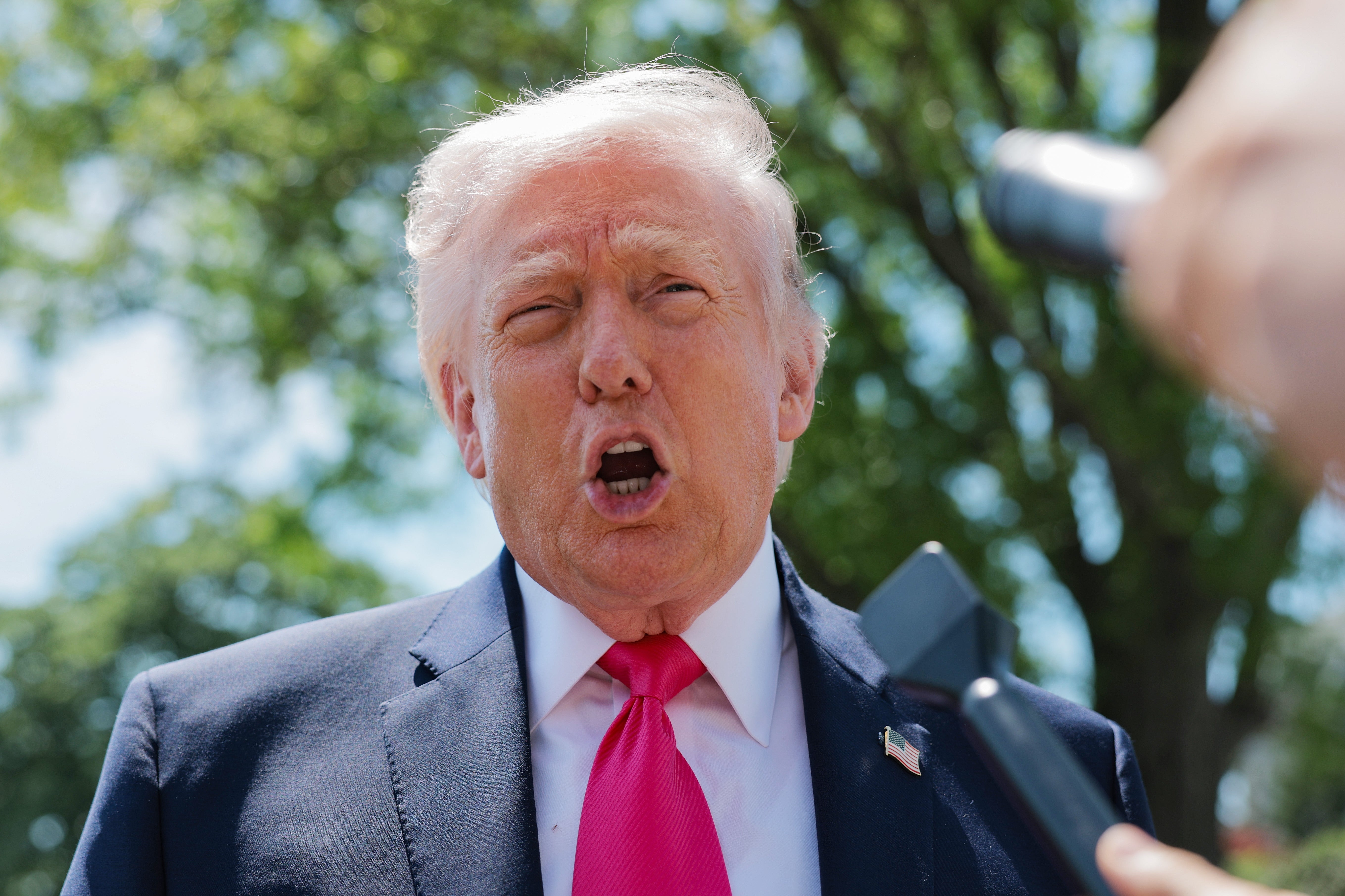 Person successful  a suit   speaking outside, with microphones held successful  beforehand   of them. Trees are disposable   successful  the background