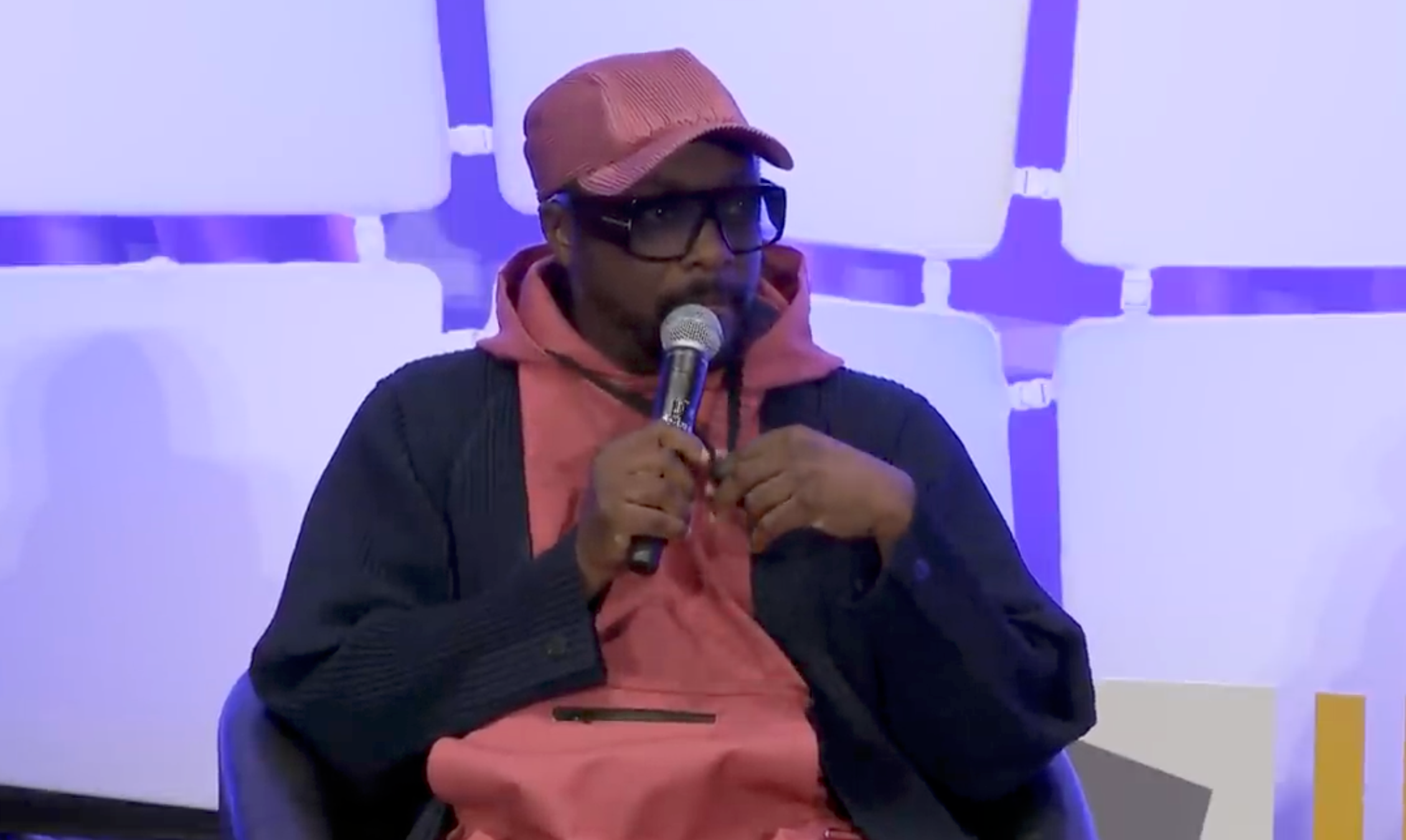 A idiosyncratic   successful  a cap, sunglasses, and casual attire speaks into a microphone portion    seated astatine  a sheet  event