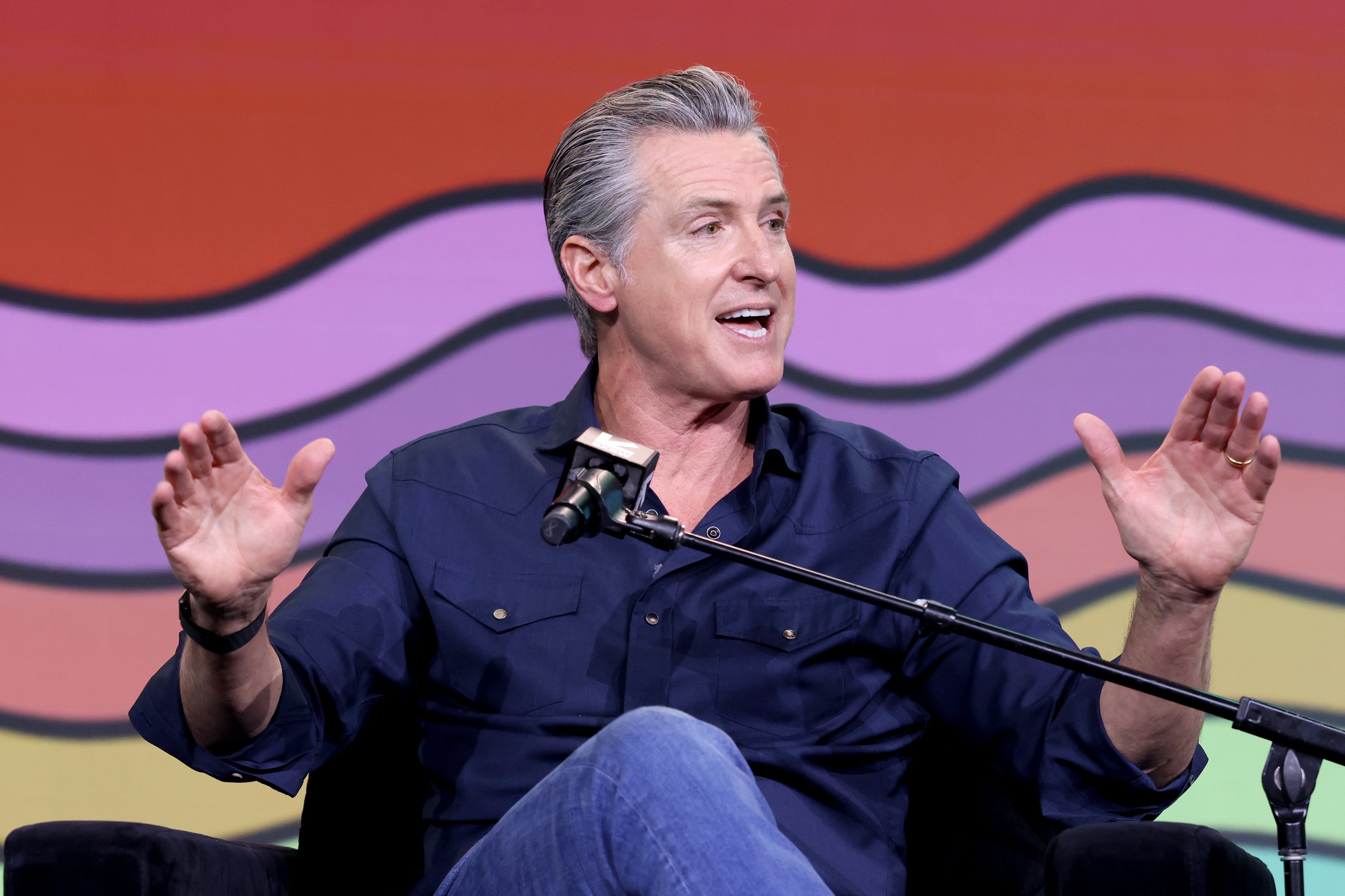 A idiosyncratic   speaking animatedly connected  stage, seated, wearing a casual button-up garment  and jeans, with a microphone successful  front