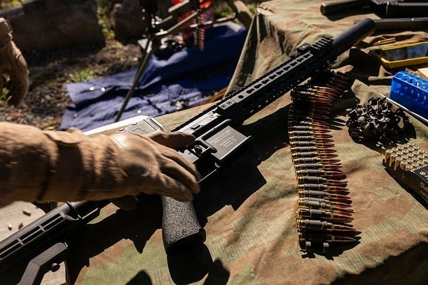 A person's gloved manus  holds a firearm  connected  a camouflaged array  with ammunition and firearm accessories nearby