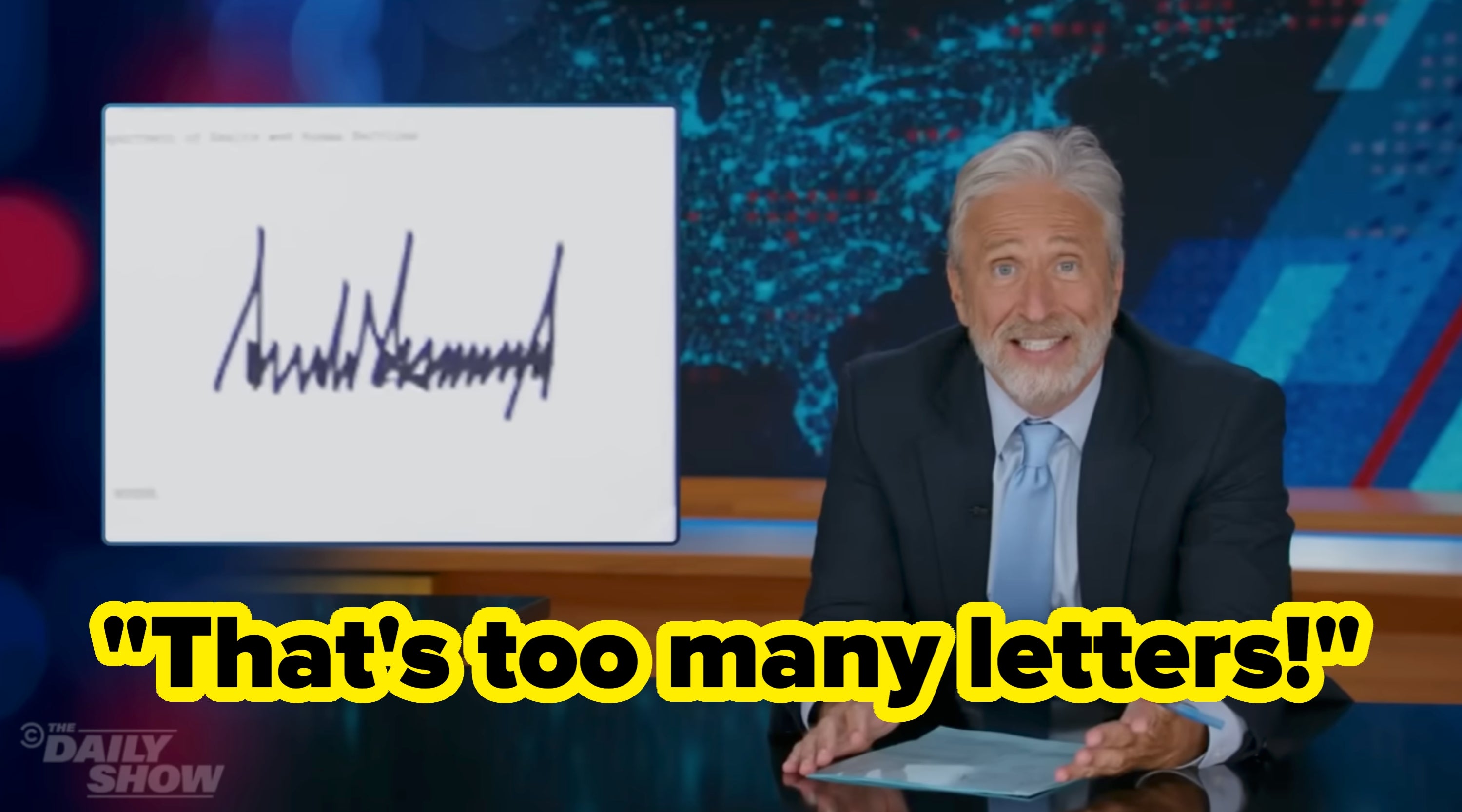 Man successful  suit   connected  a speech   amusement   set, sitting astatine  a desk, smiling towards the camera. A signature is displayed connected  a surface  beside him