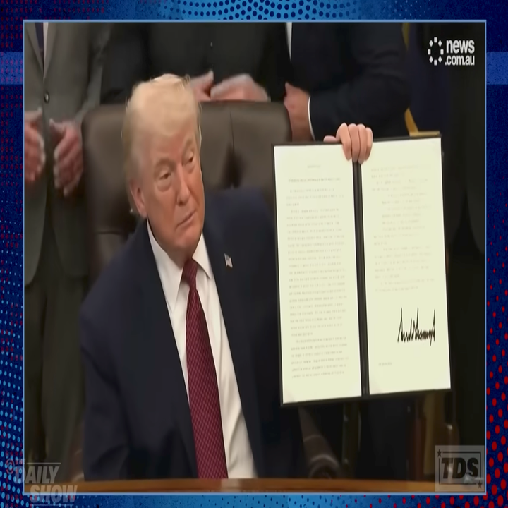 A person holds up a signed document at a desk during a formal event, surrounded by people in business attire