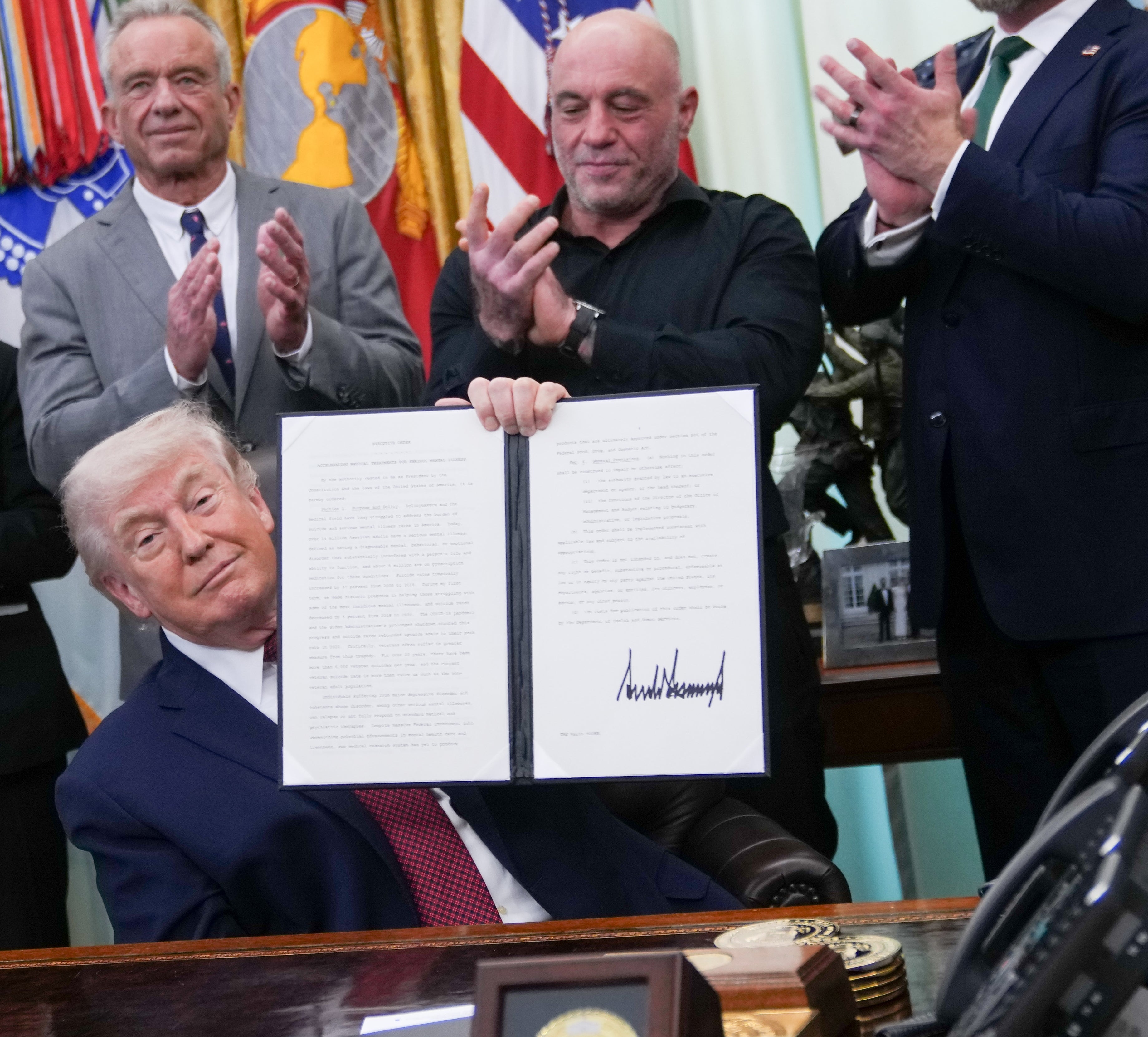 A radical  of men successful  suits clapping arsenic  Donald Trump seated holds up   a signed papers  successful  an authoritative  mounting  with flags successful  the background