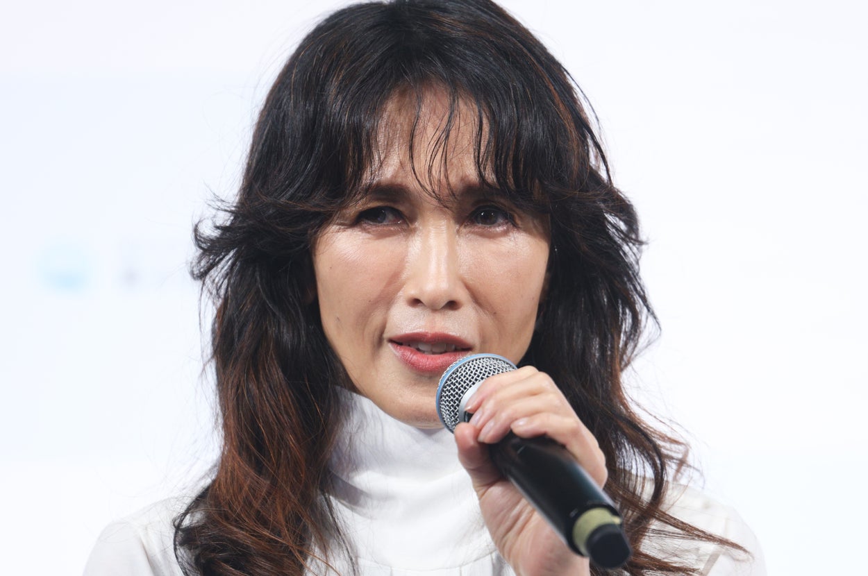 女性がマイクを持って話している様子。形式的な場面で、白いトップスを着用している。