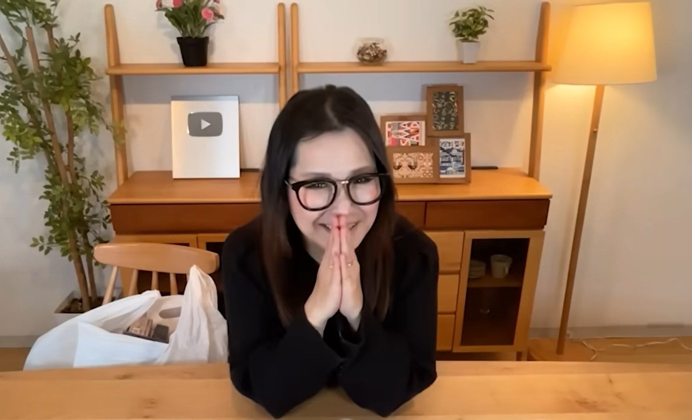 女性が室内で手を合わせて笑顔。背後には棚に植物や飾りが置かれている。