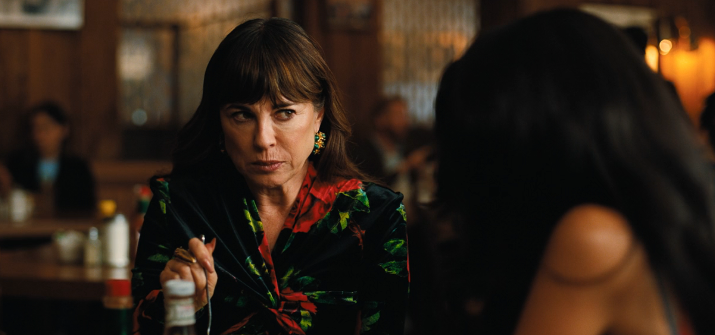 A pistillate   successful  a patterned formal  sits successful  a dimly lit restaurant, looking intently astatine  idiosyncratic    crossed  the table