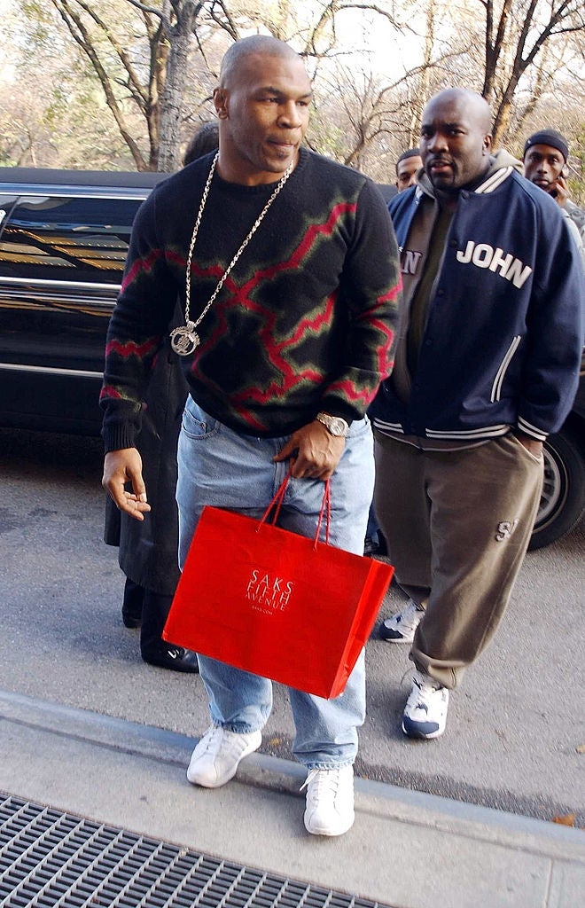 Person successful  a casual achromatic  and reddish  sweater with a concatenation  necklace, carrying a buying  bag, walking connected  a sidewalk. Another idiosyncratic   follows behind