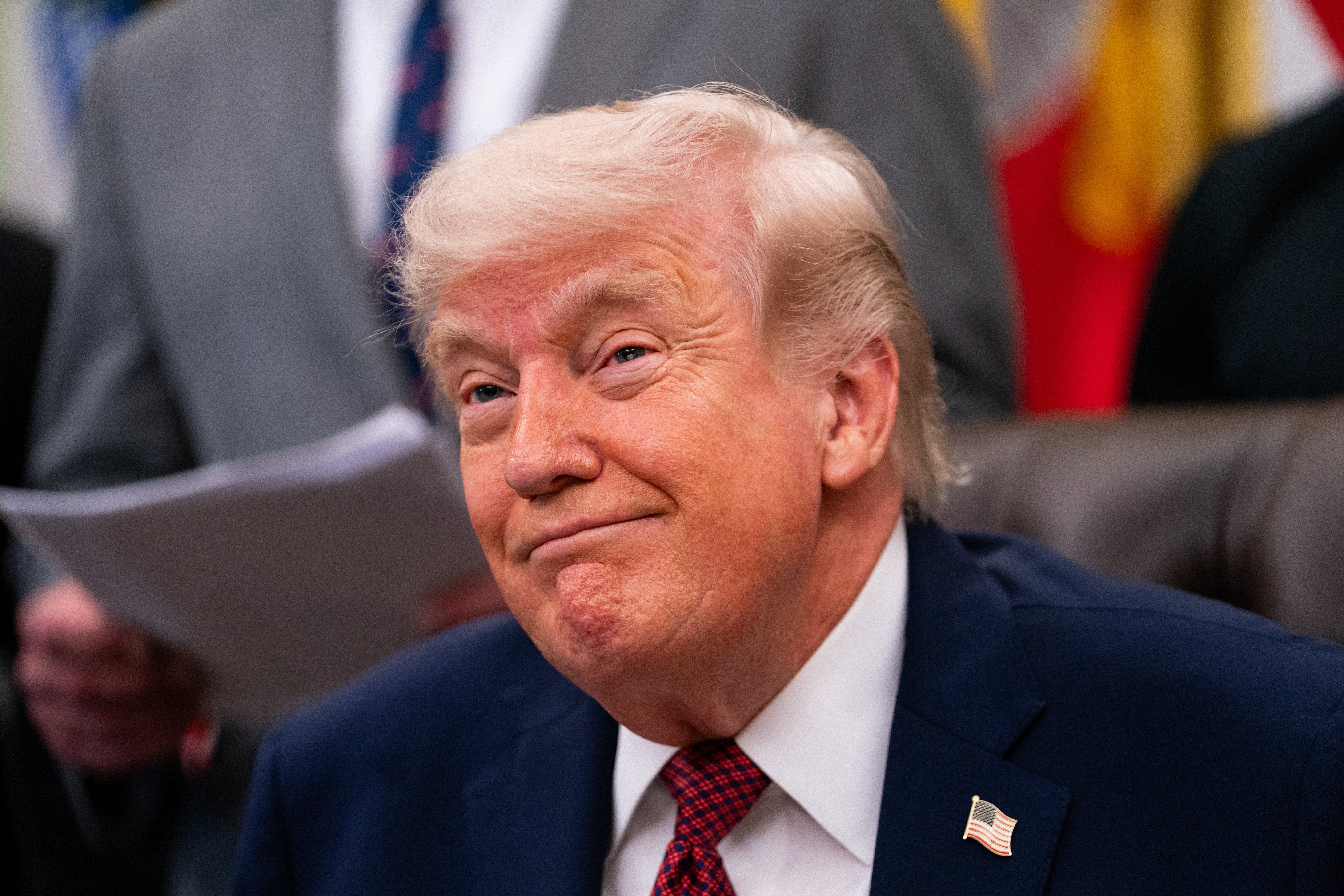 A idiosyncratic   successful  a suit   and necktie  sits astatine  a desk, looking somewhat  upwards with a half-smile; others look  somewhat  blurred successful  the background
