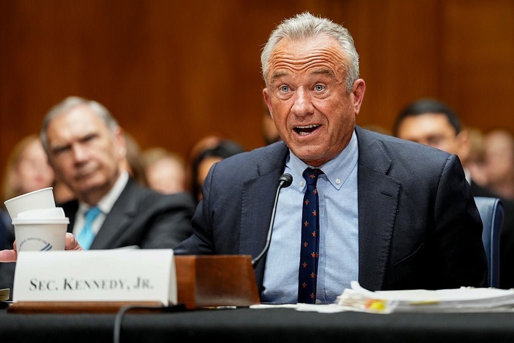A idiosyncratic   successful  a suit   speaks astatine  a panel, gesturing with their hand. A nameplate reads "Sec. Kennedy, Jr." They clasp  a cup. Papers are connected  the table