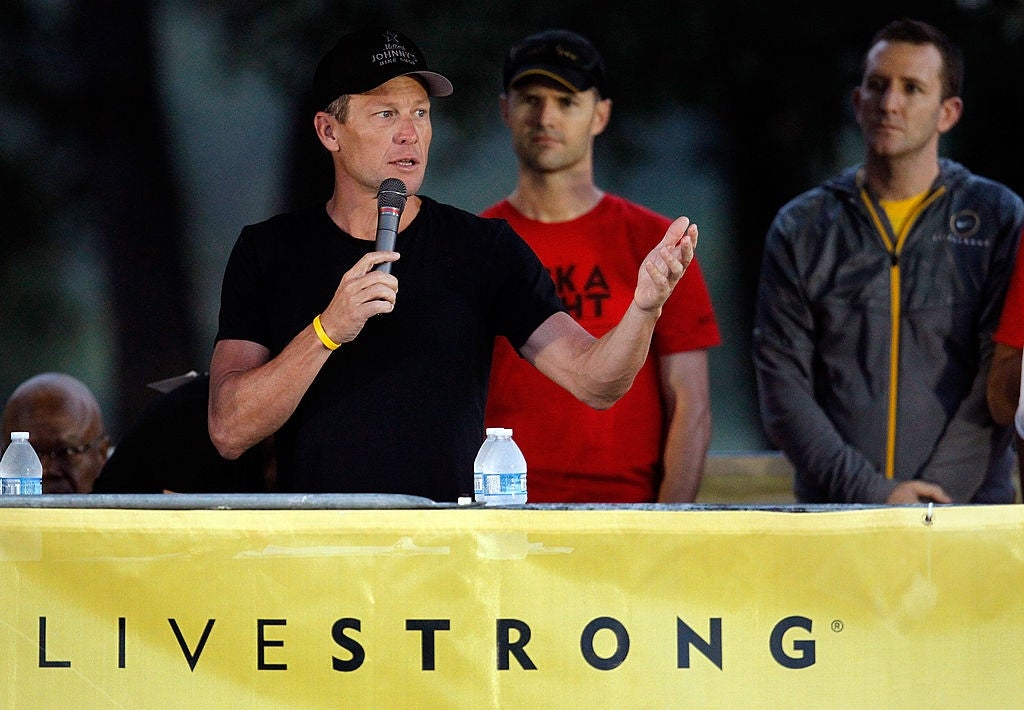 A idiosyncratic   successful  casual attire speaks astatine  a podium with a microphone, flanked by others, successful  beforehand   of a banner that says "LIVESTRONG."