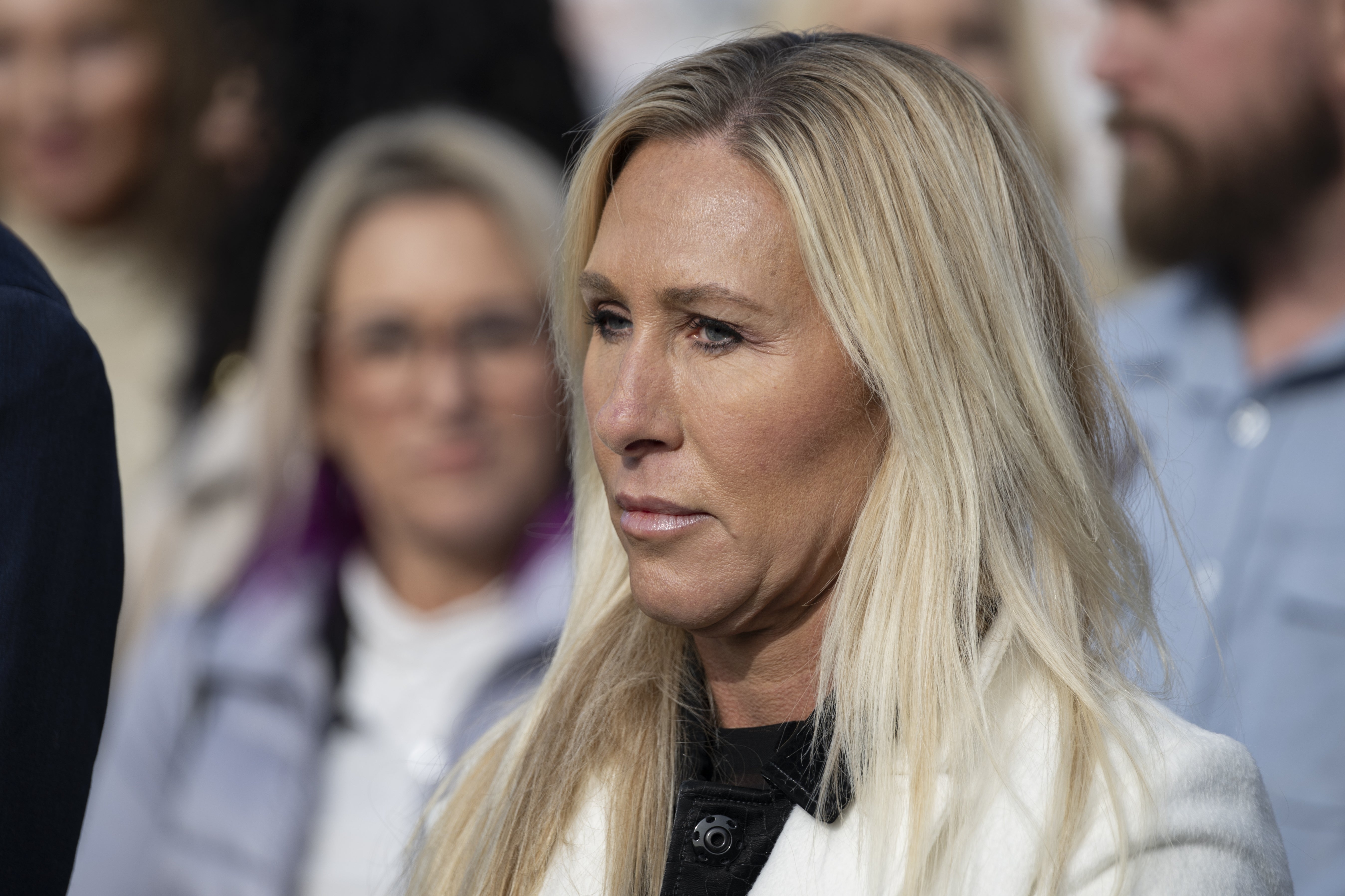 En person med langt blondt hår i en formell setting, iført en lys frakk. Andre mennesker er uskarpe i bakgrunnen