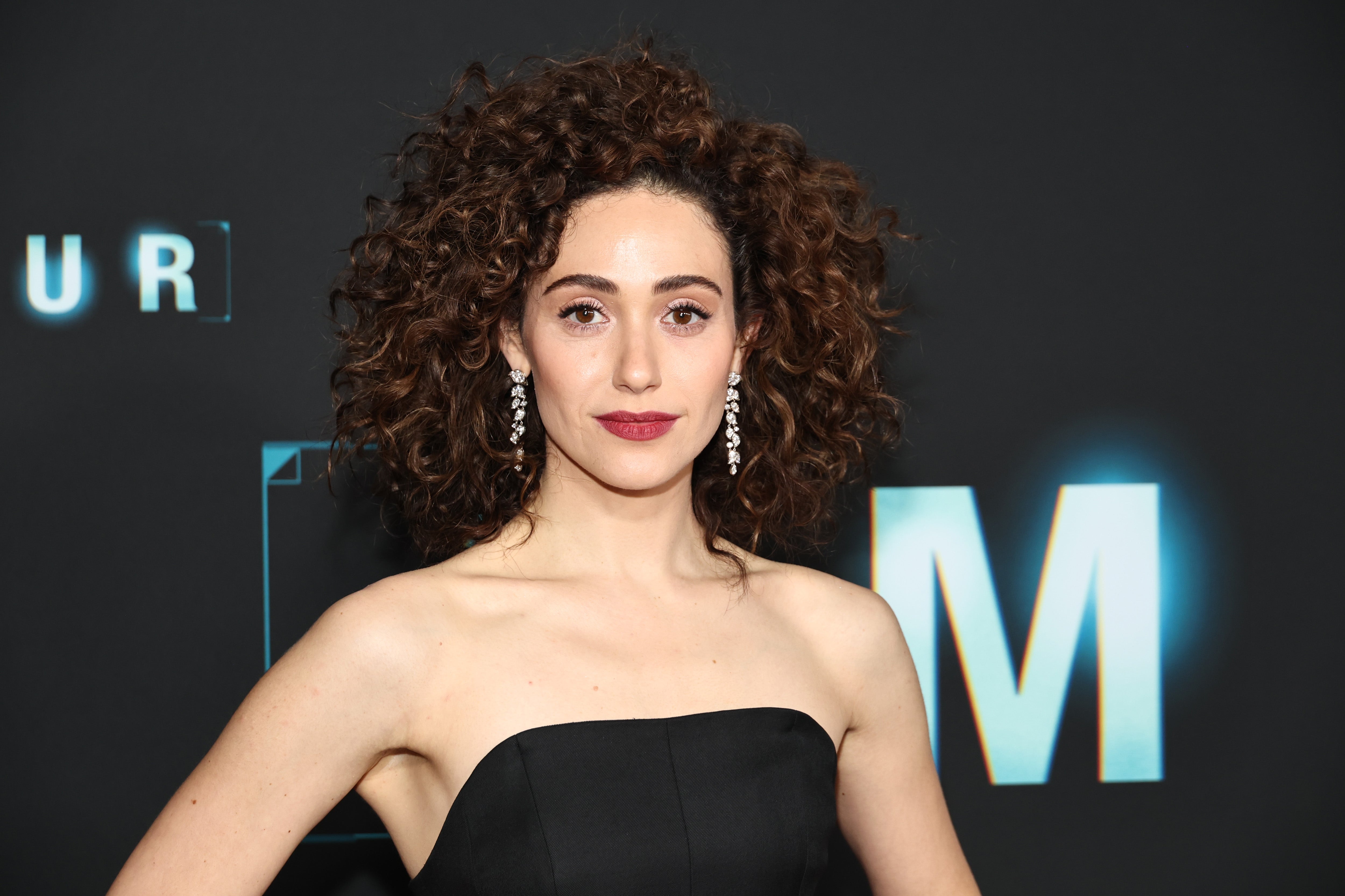 Emmy Rossum with curly hairsbreadth  successful  a strapless formal  stands successful  beforehand   of a backdrop with text