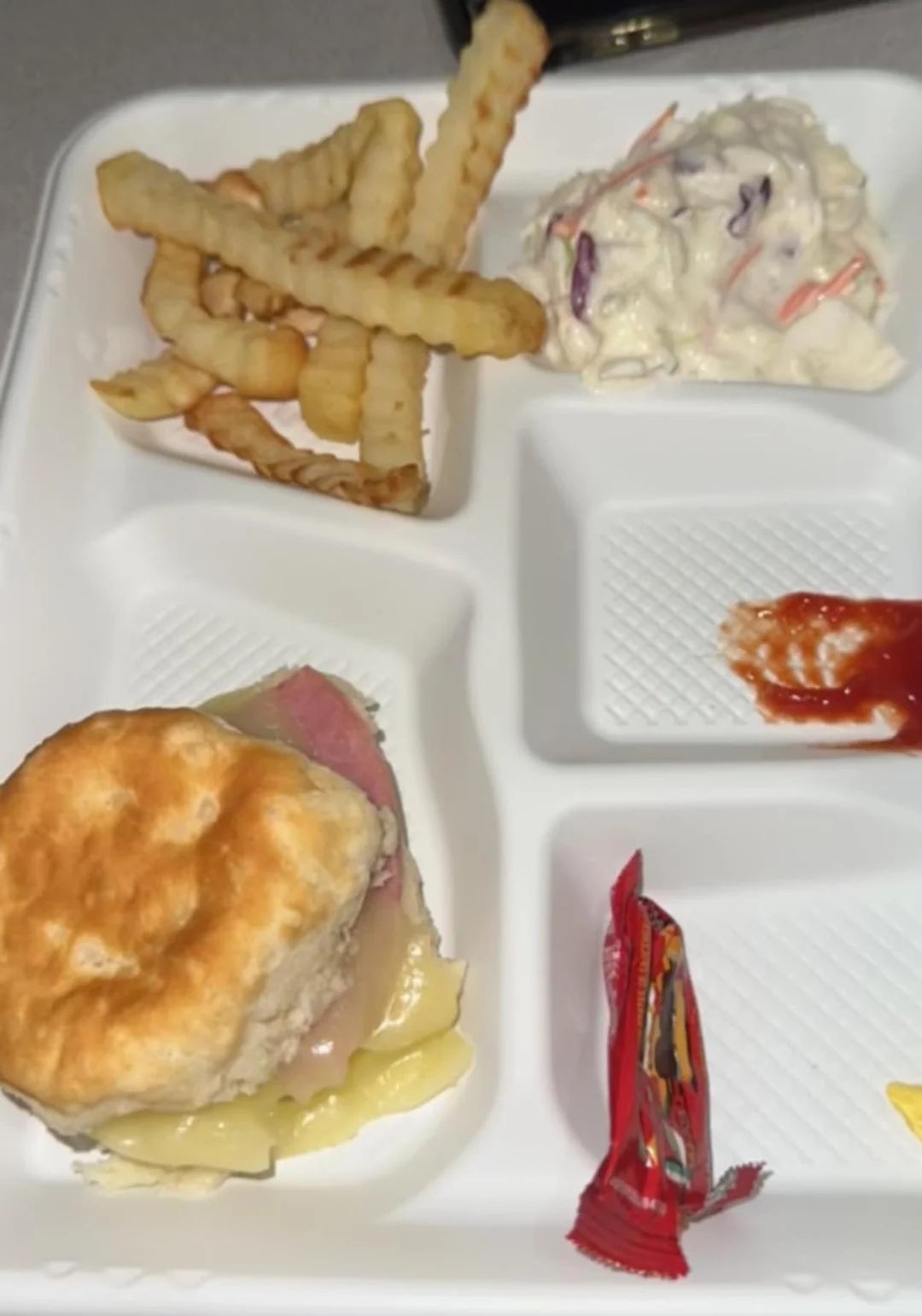 Container with crinkle-cut fries, coleslaw, biscuit sandwich with ham and cheese, ketchup, and a tiny  condiment  packet