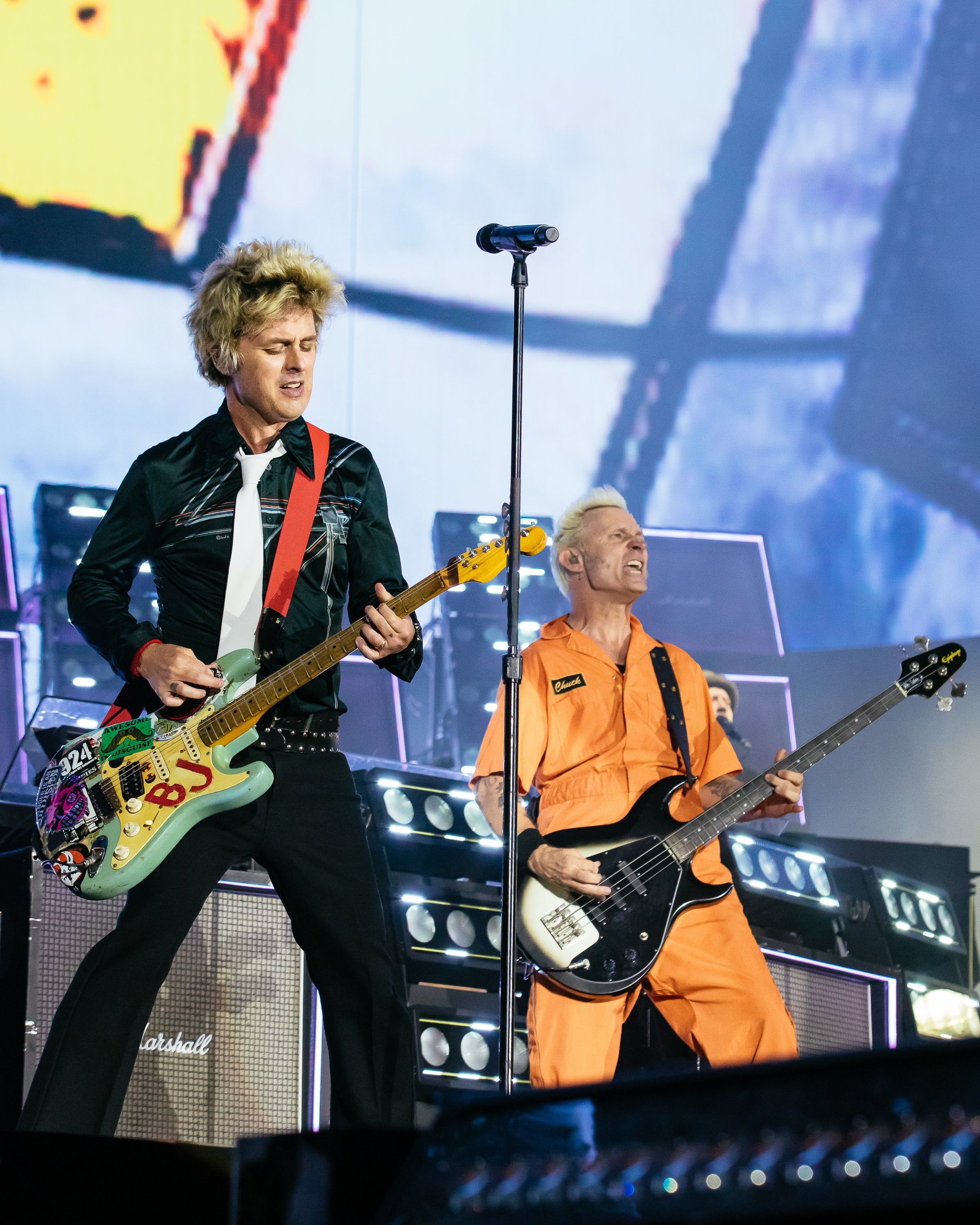 Two musicians execute  energetically connected  stage, 1  wearing a stylish garment  and tie, the different   successful  a jumpsuit with a guitar and bass, respectively