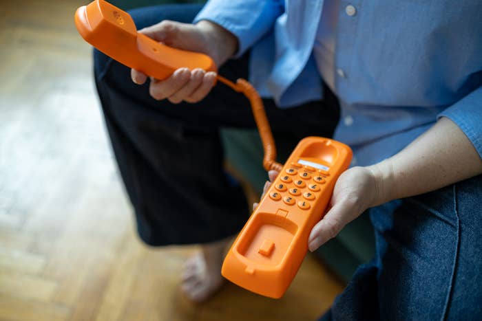 Person holding a retro-style orangish  corded telephone   with some  hands, wearing bluish  garment  and acheronian  pants