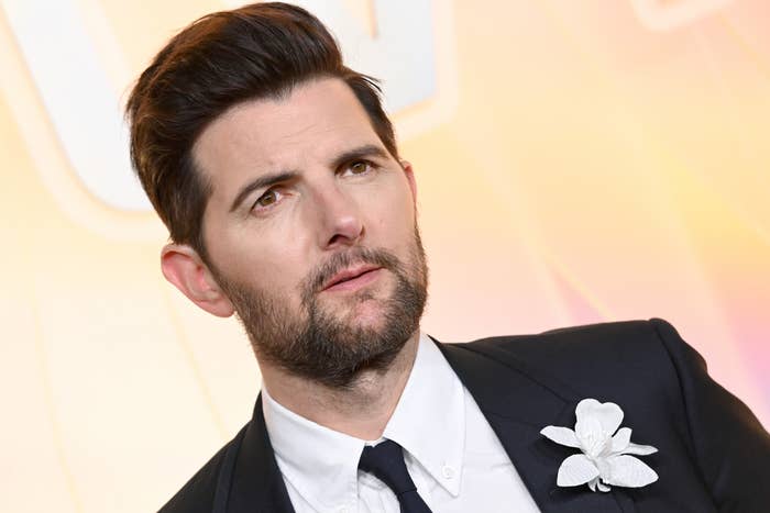Adam Scott on a red carpet wearing a suit with a floral pin, looking slightly to the side