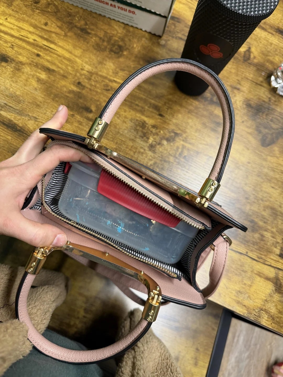 Hand holding an unfastened  handbag, revealing an organized interior with a wide   container  inside, connected  a woody  tabletop