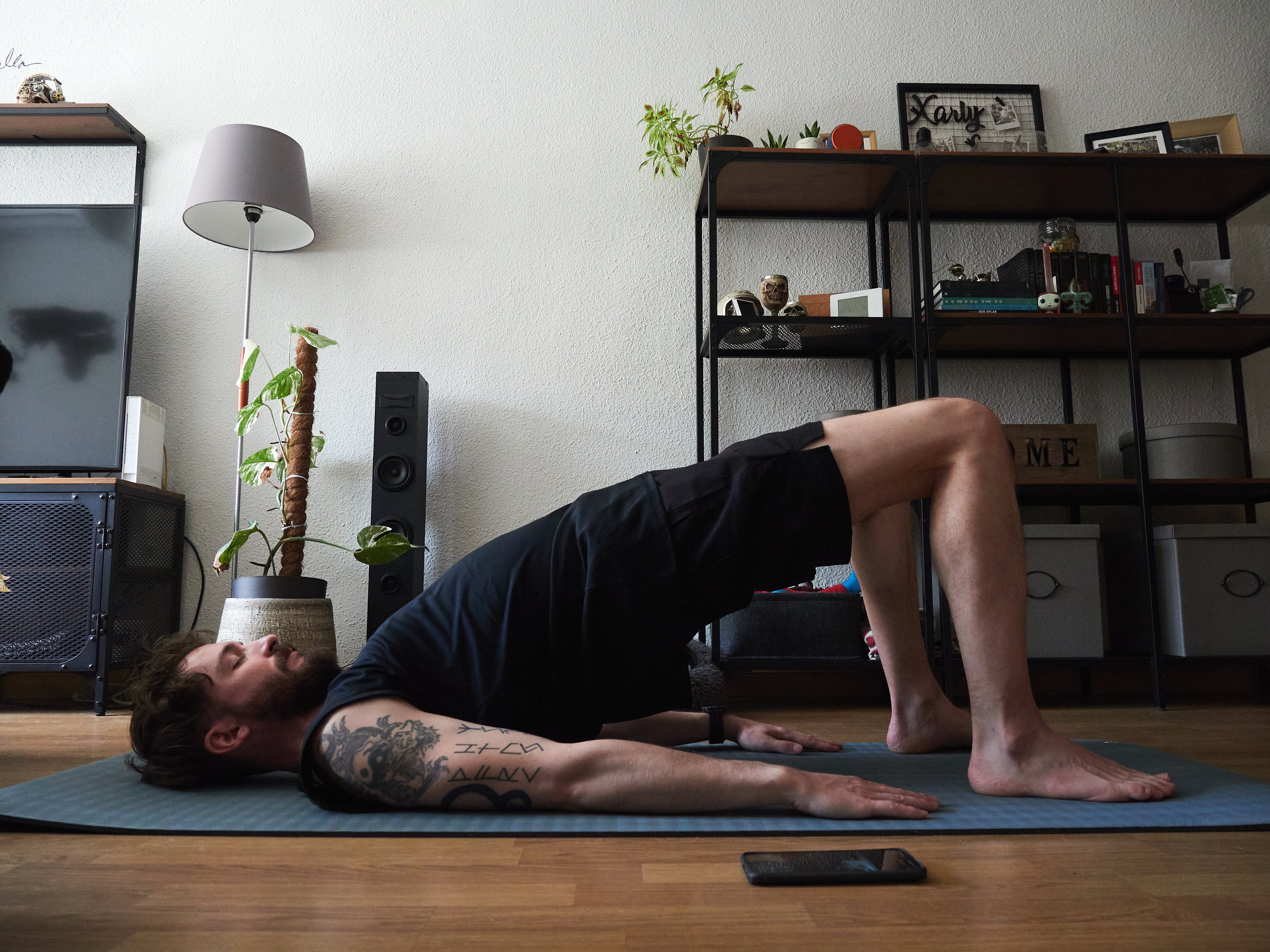 En person med tatoveringer gjør en brostilling på en yogamatte i en moderne stue med hyller og en plante
