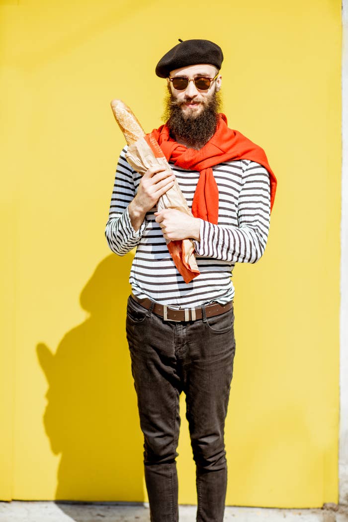 A antheral   with a beard and sunglasses holds a baguette. He's wearing a beret, striped shirt, and reddish  scarf, lasting  successful  beforehand   of a plain background
