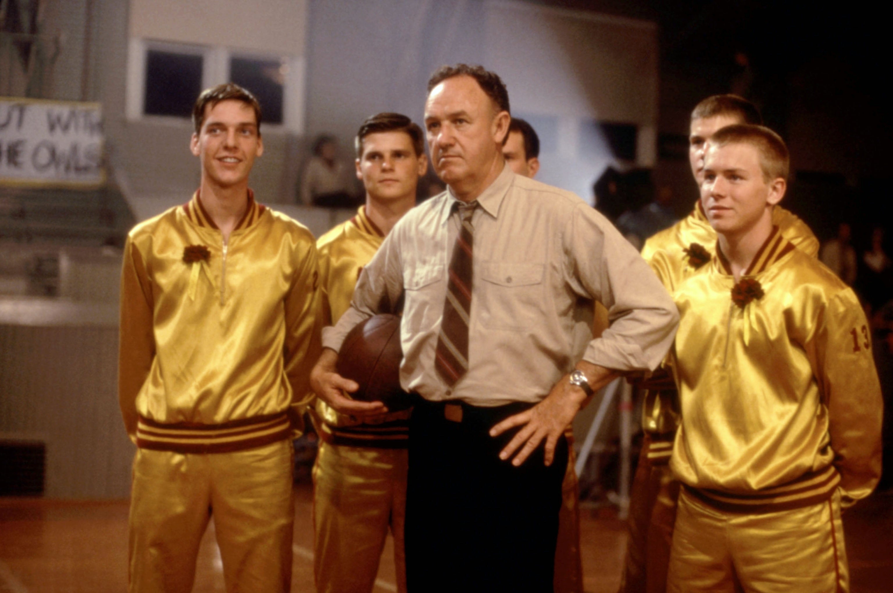 A basketball coach stands confidently with his team wearing matching uniforms, holding a basketball, in a sports setting