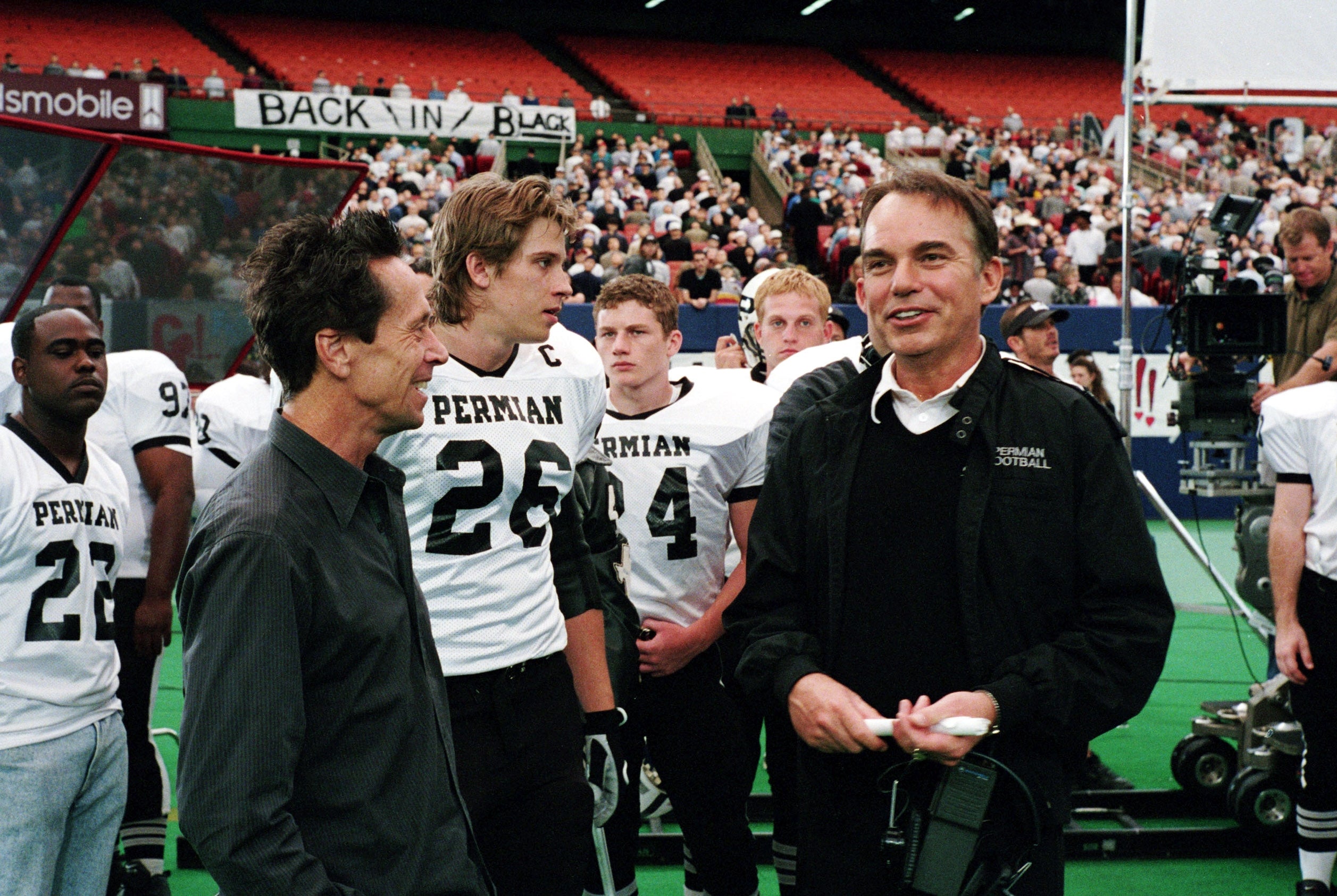 Actors and crew in football uniforms on a sports field. Film equipment visible in the background