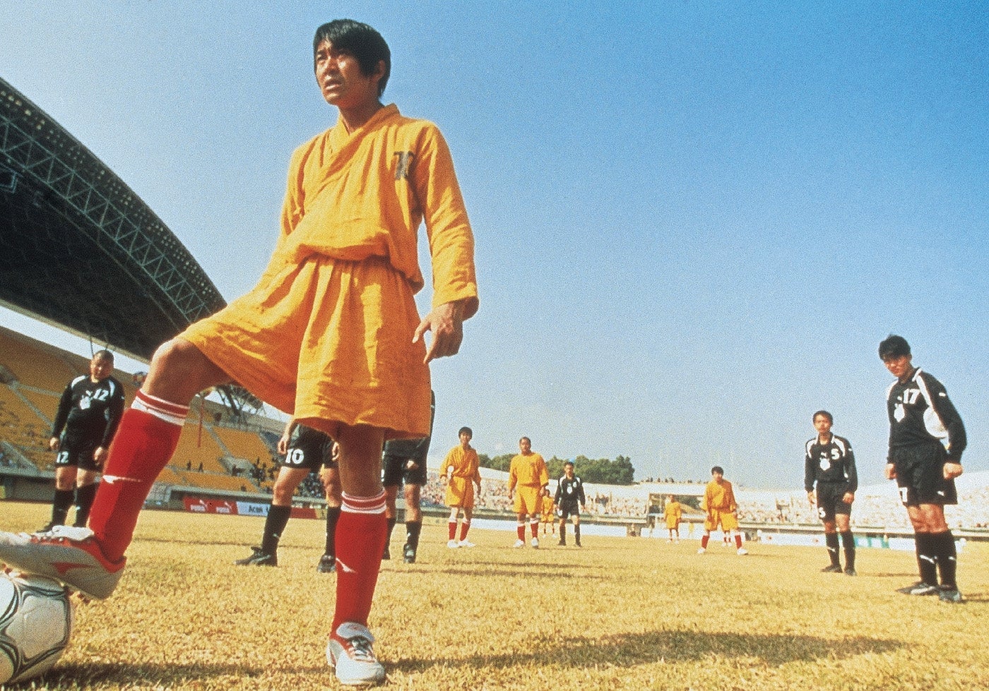 A soccer player in a robe readies to kick the ball on a field, surrounded by teammates and opponents