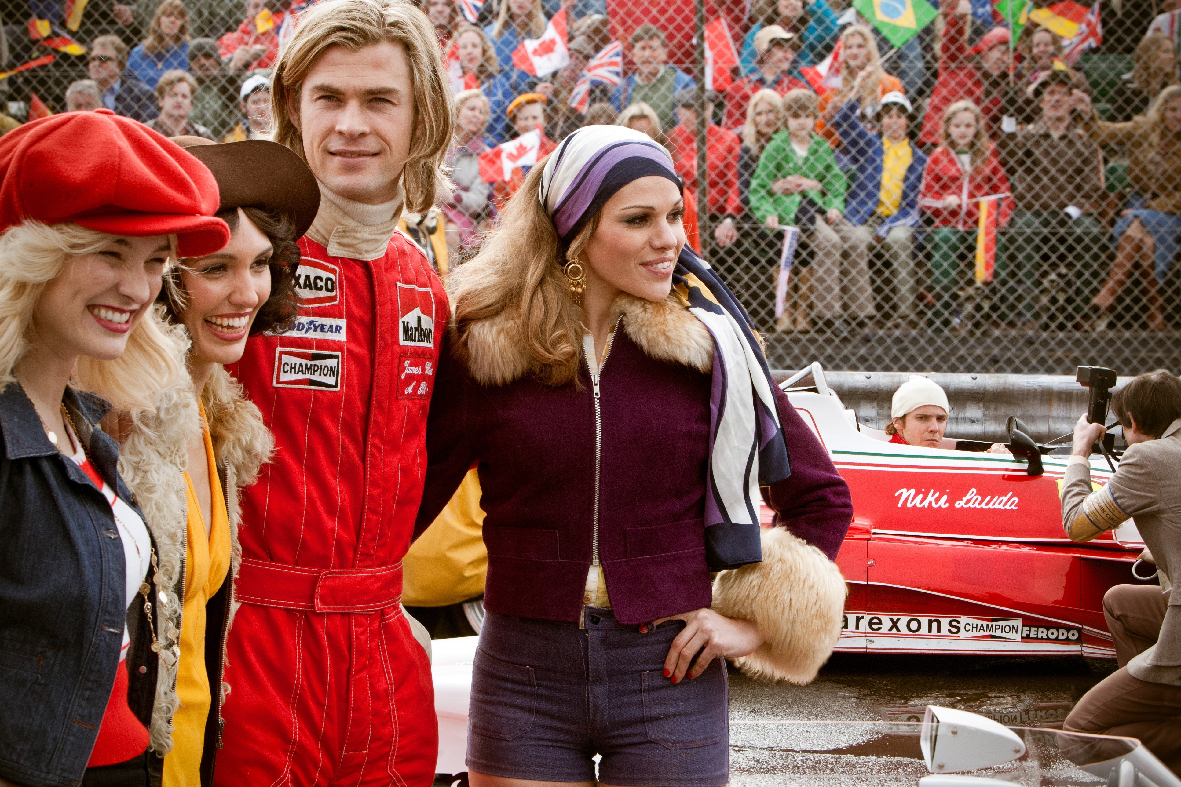A group of people in vintage racing attire pose at a racetrack, with one person in a red racing suit. A vintage race car is visible in the background