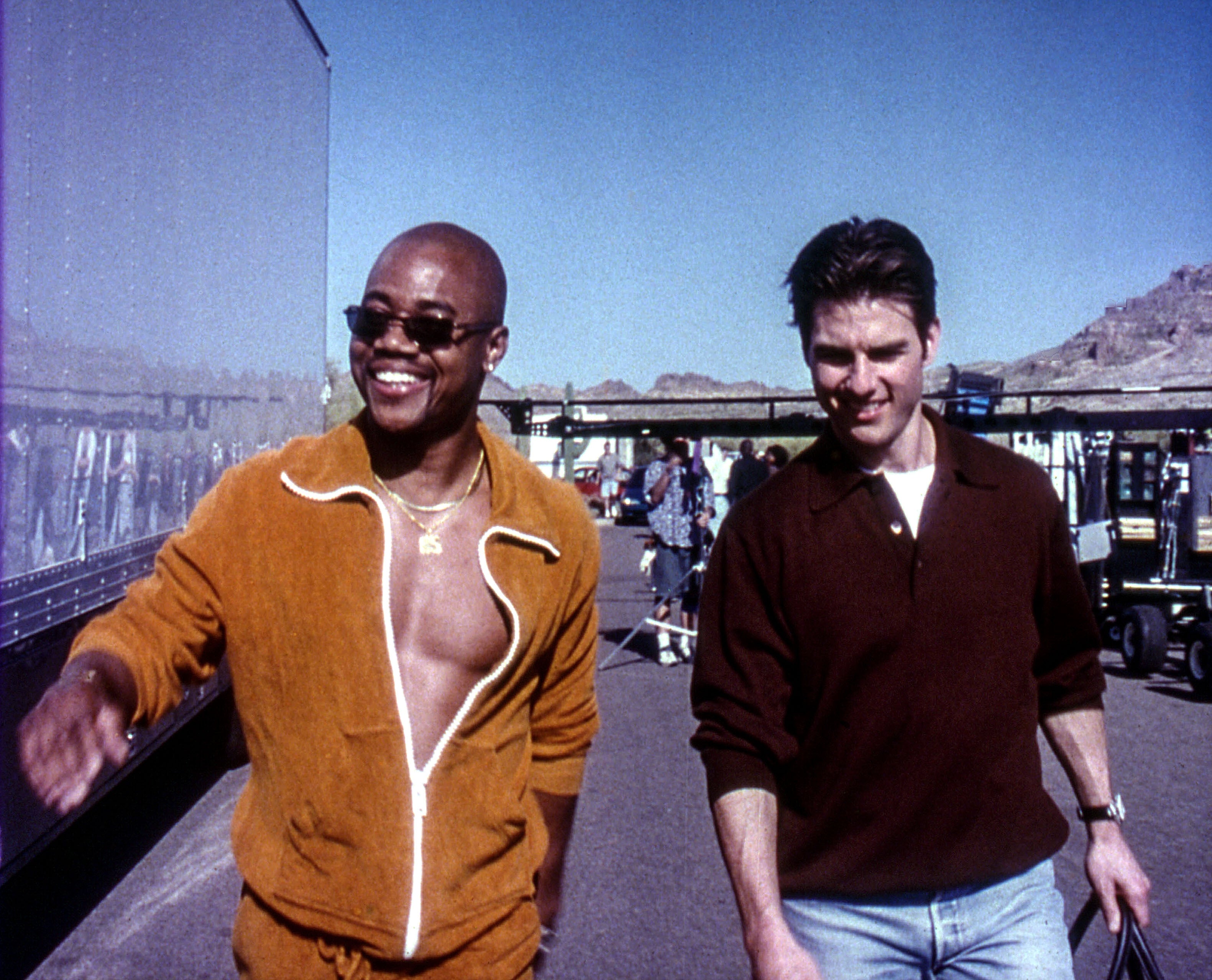 Two people walking outdoors, one in an open jacket and sunglasses, the other in a sweater, both smiling. Appears casual and informal