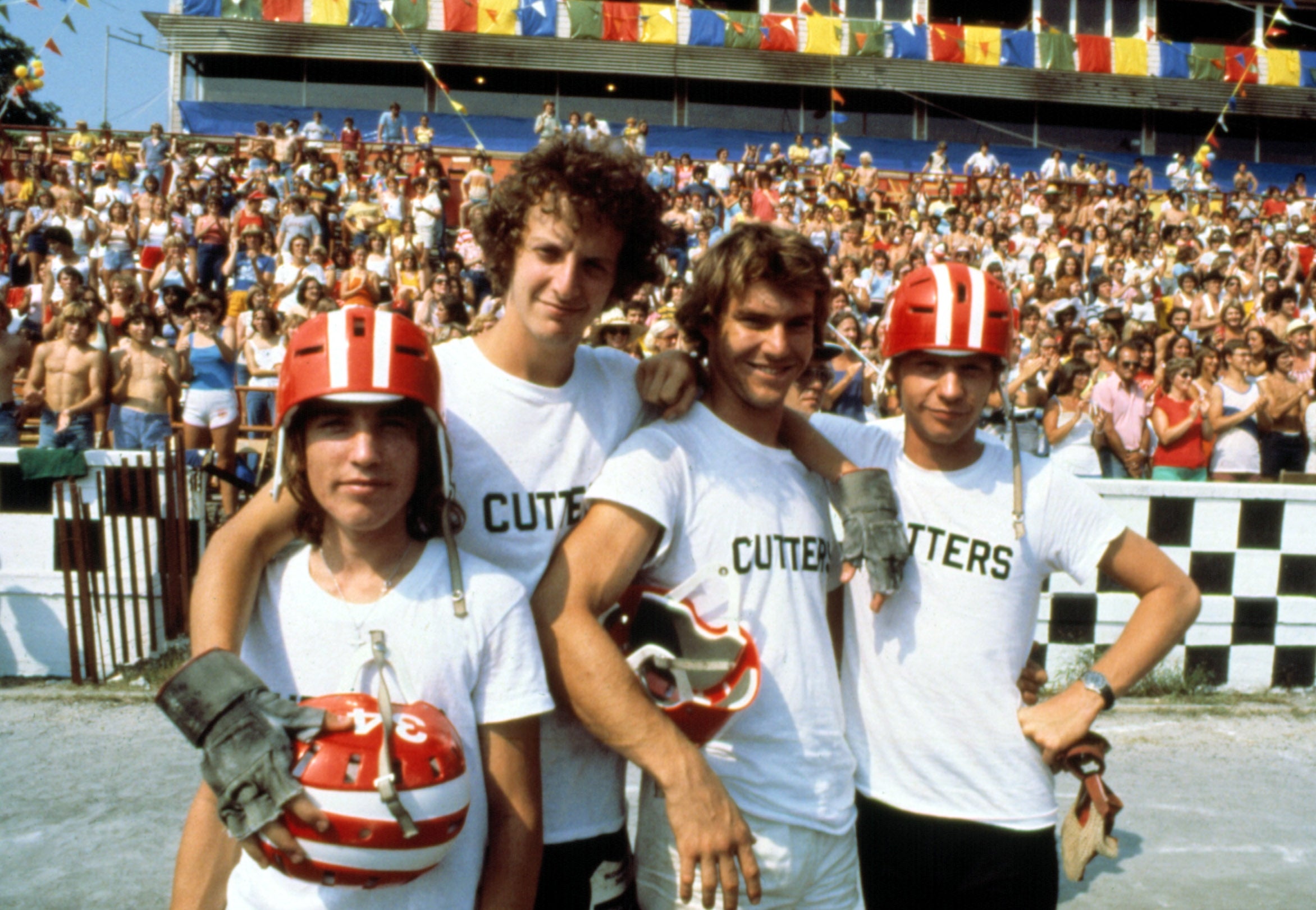 Four people in "Cutters" shirts and helmets stand together on a racetrack, with a cheering audience in the background