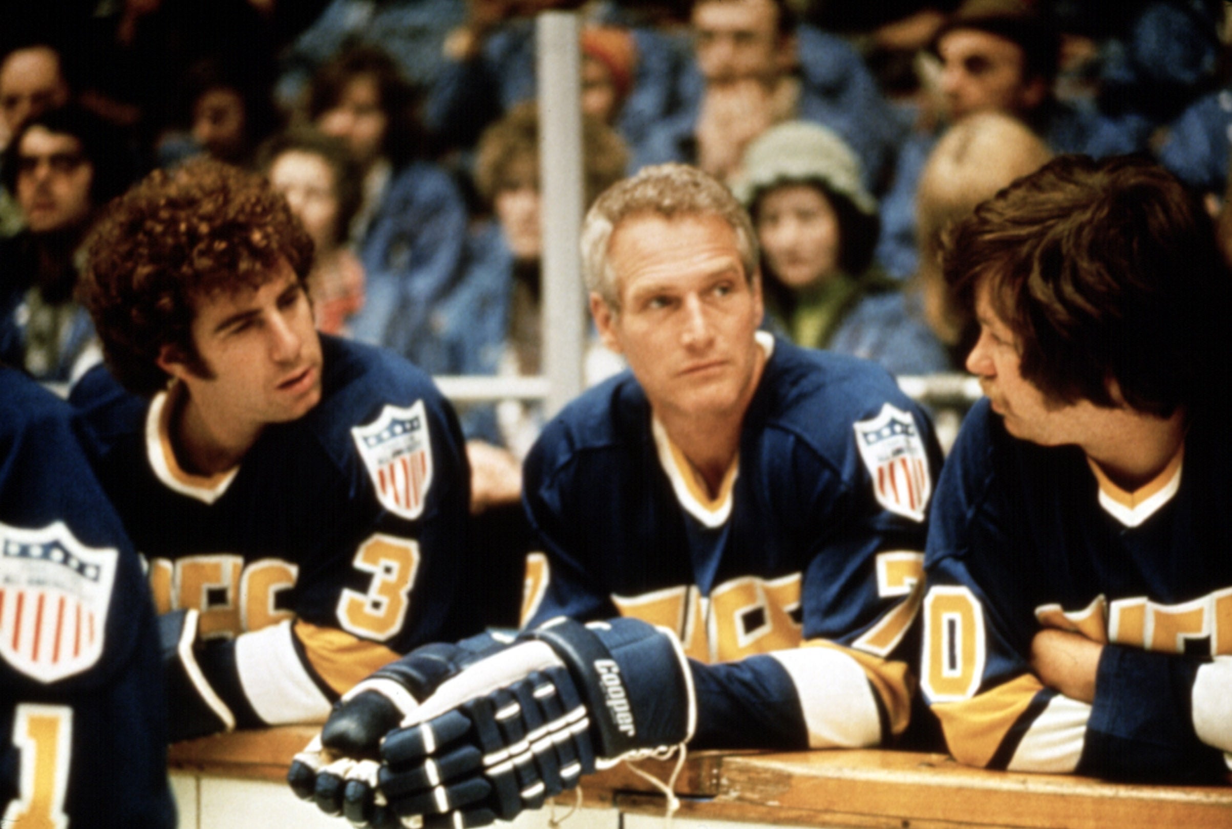 Three actors in hockey jerseys sit on a bench, appearing focused and engaged in conversation