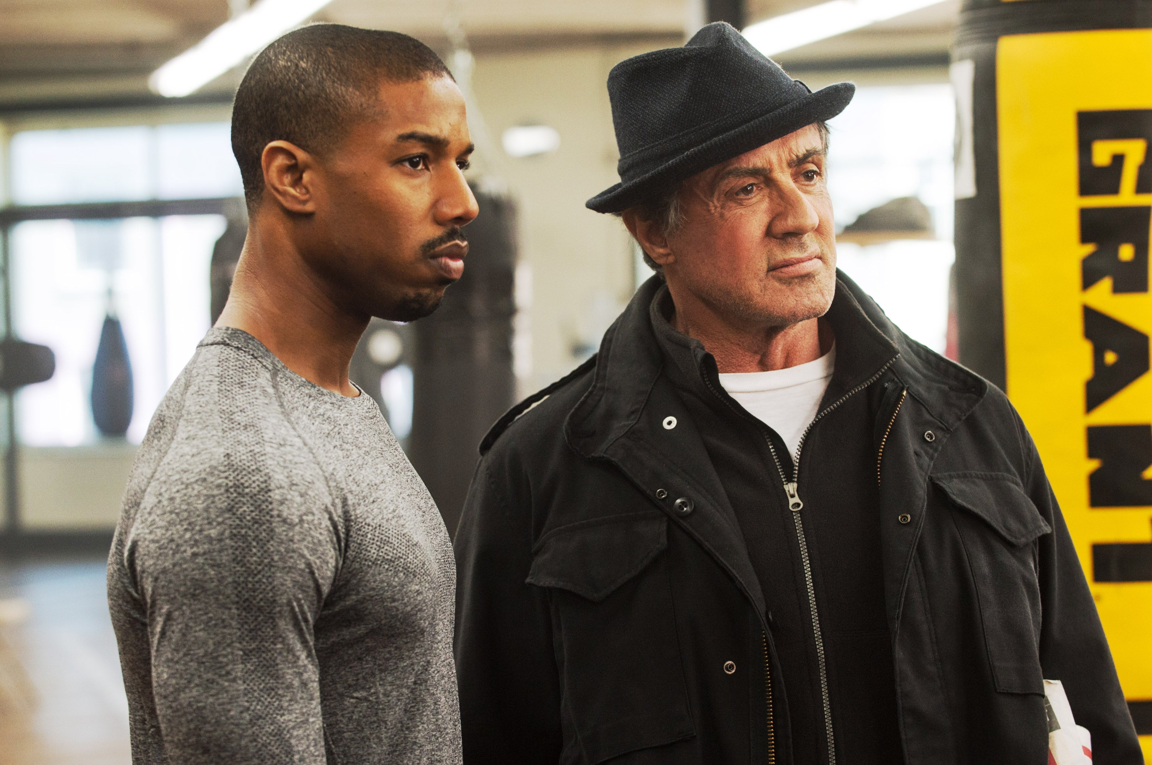 Two actors in a gym scene, one in a fitted shirt and the other in a jacket and fedora, looking focused and determined