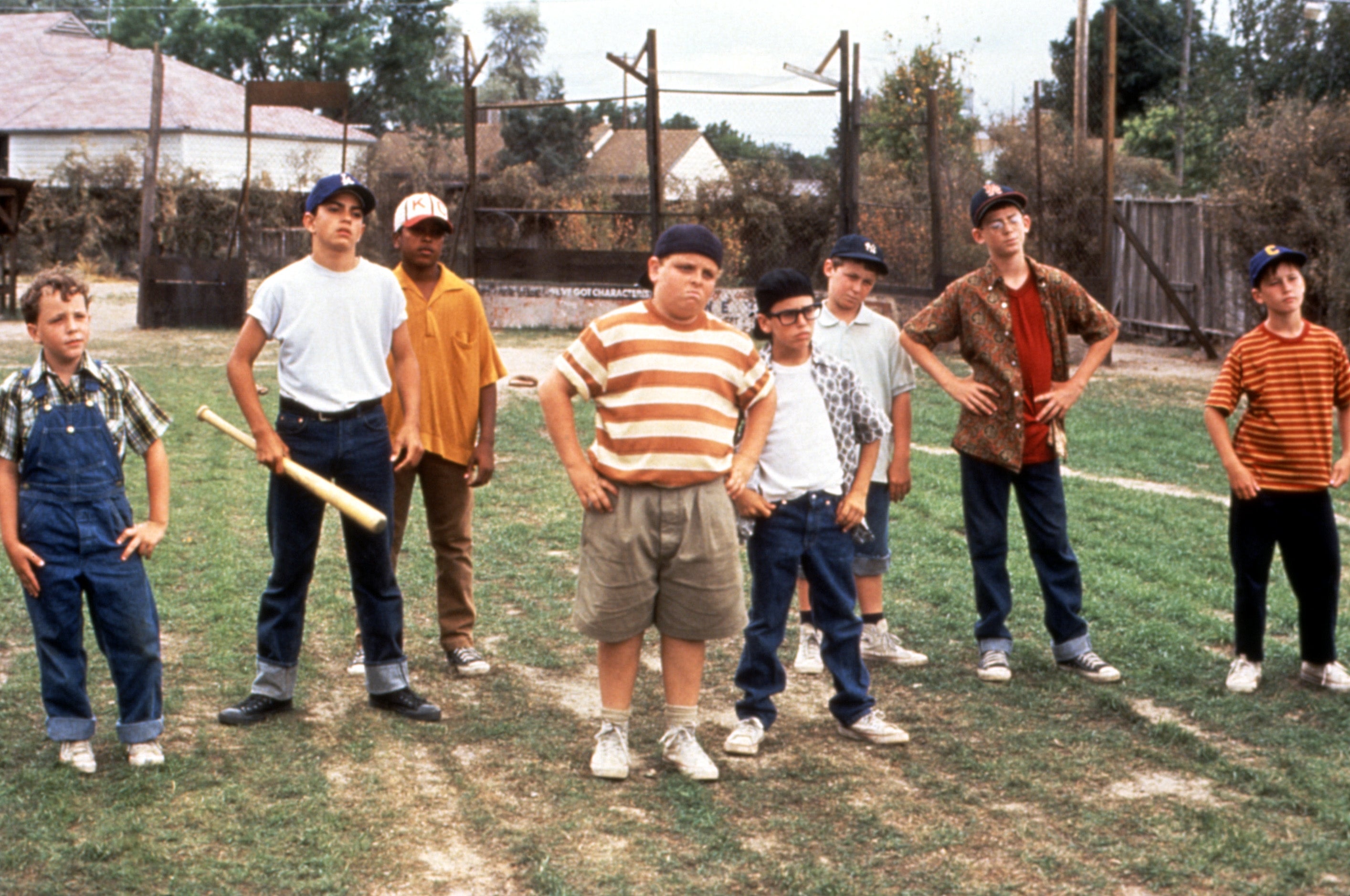 A group of kids on a baseball field stand confidently, wearing casual baseball attire, with one holding a bat