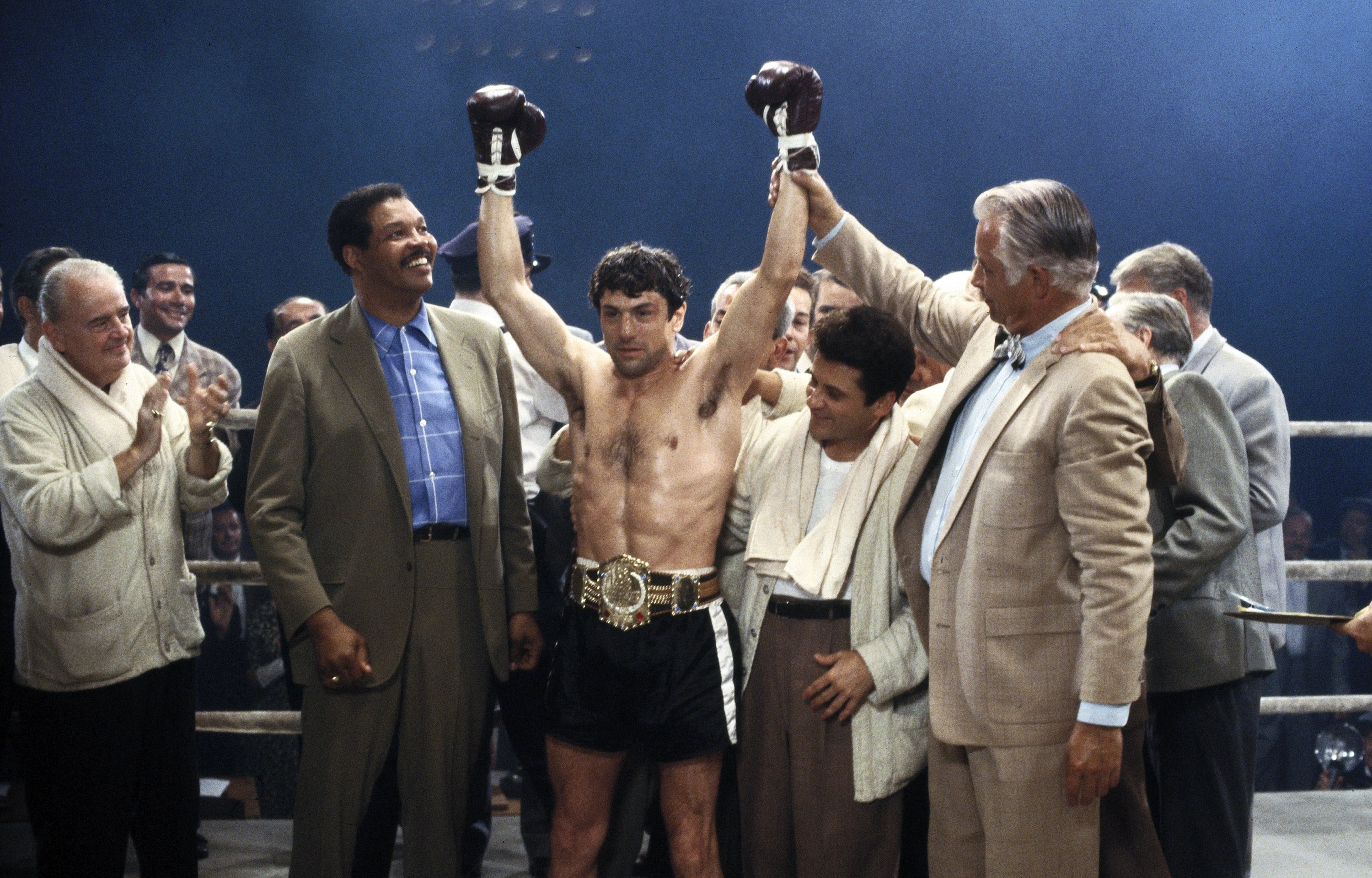 A victorious boxer stands in the ring with arms raised, surrounded by supporters and officials in suits, celebrating a boxing match win