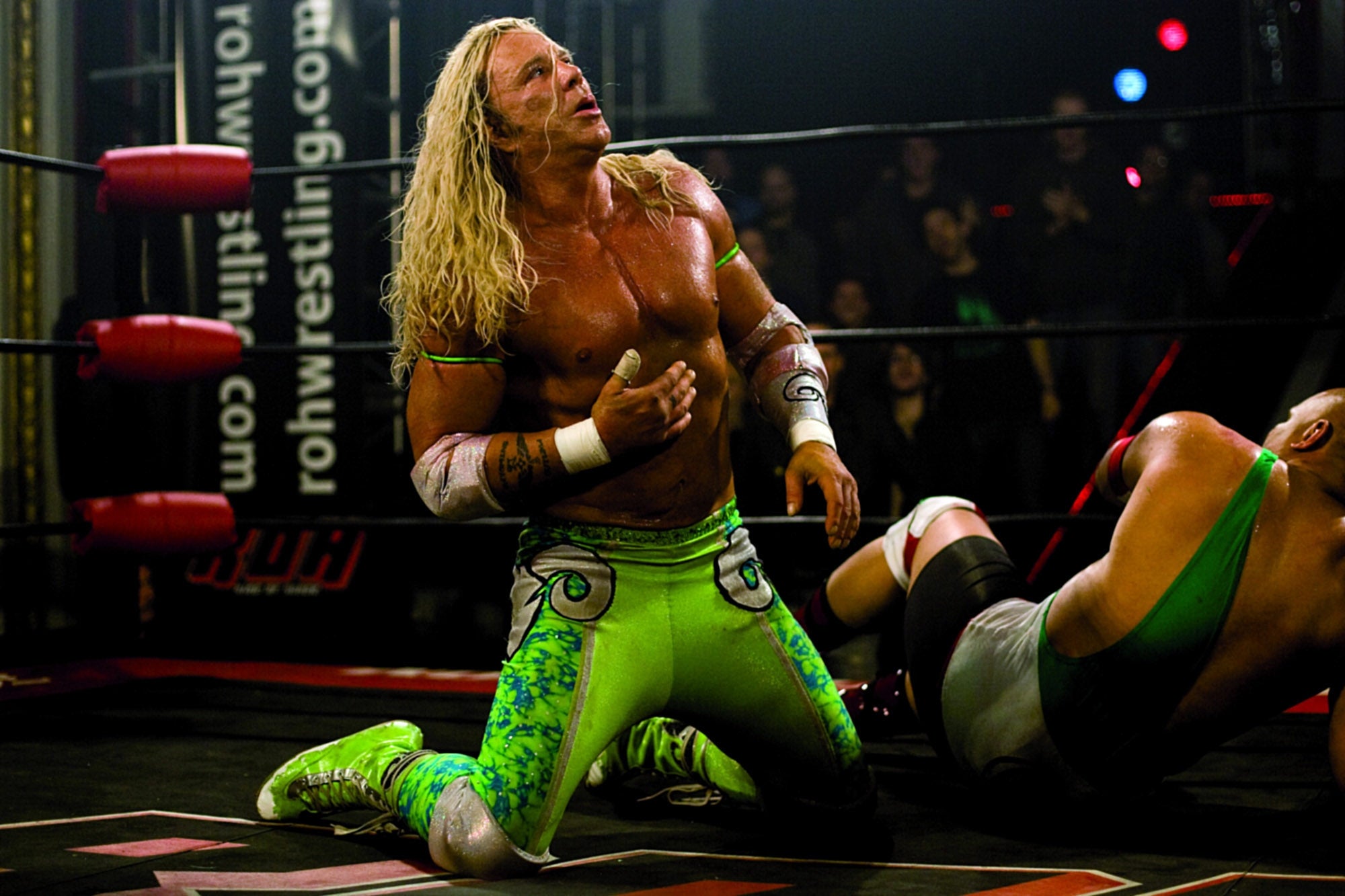 Wrestler kneeling in the ring, wearing vibrant green tights and wristbands, with long, curly hair. Another wrestler lies on the mat nearby