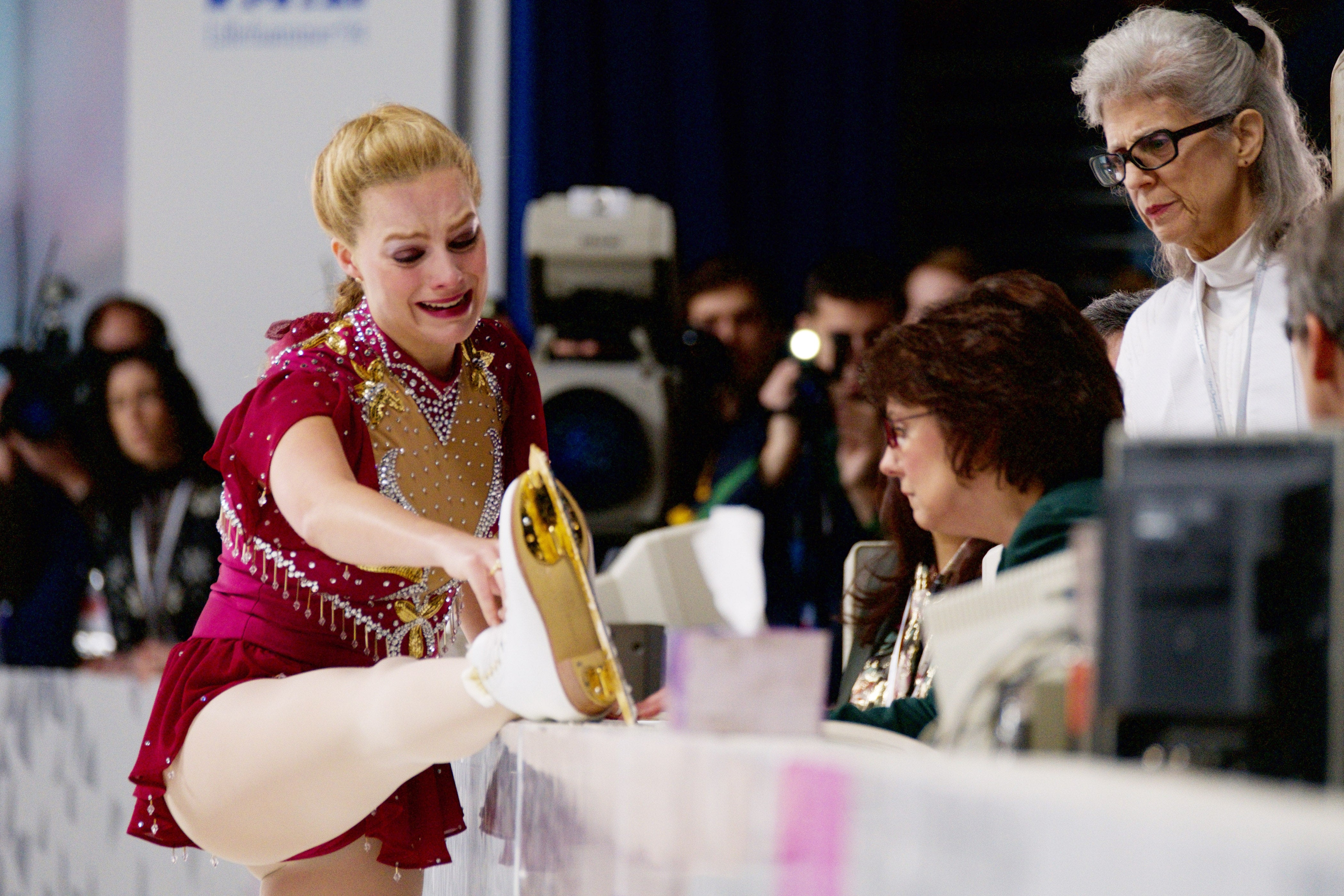 Figure skater in a maroon costume appears upset as she shows her skate to judges during a competition. Observers watch closely