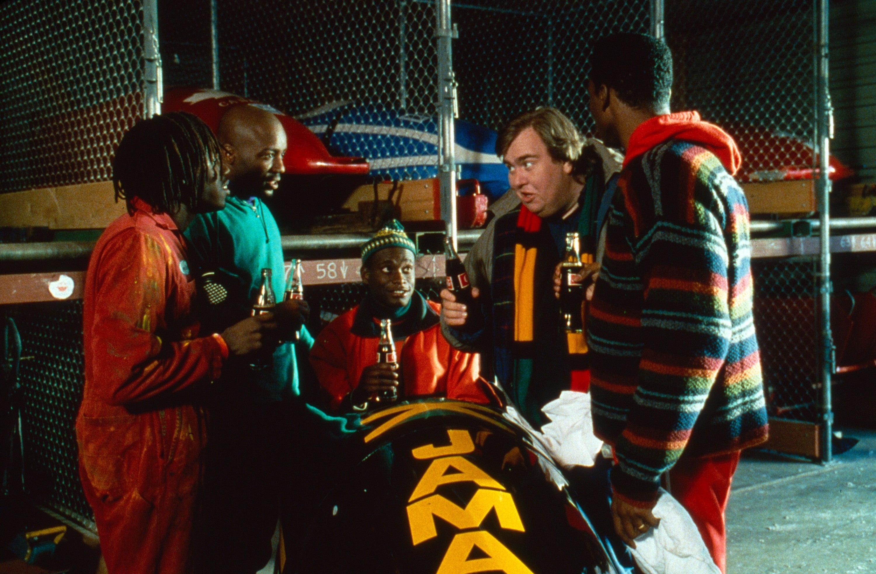 A comedic scene featuring four men in casual winter clothing gathered around a Jamaican bobsled, holding bottles, in a storage area