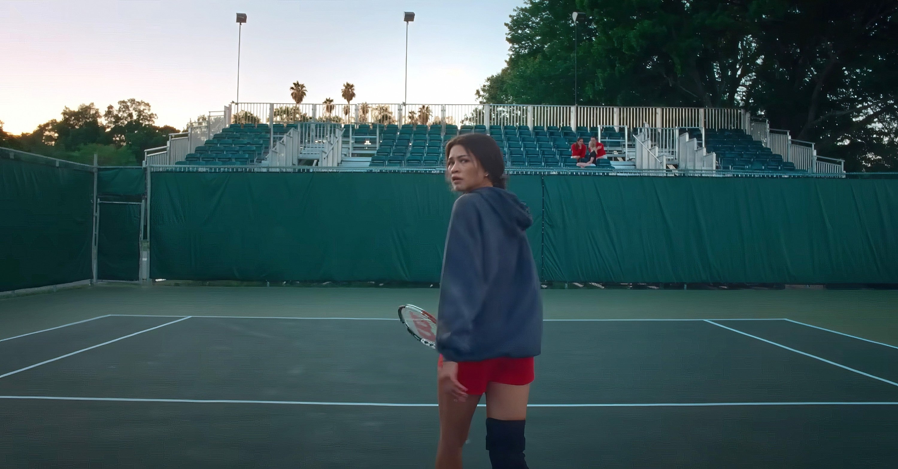 A person stands on an empty tennis court with a racket, glancing back. They wear a hoodie, red shorts, and a knee brace