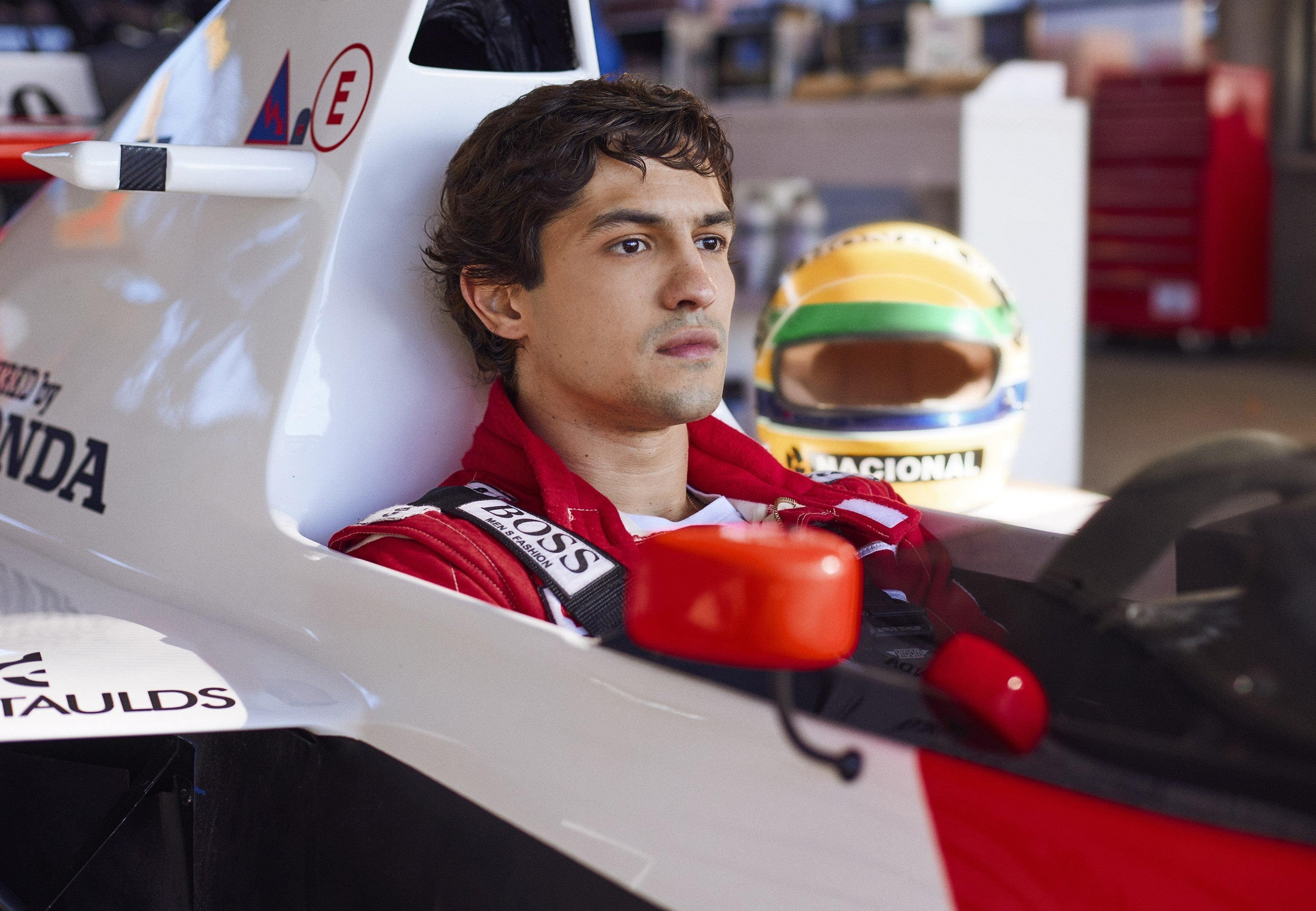 Person sitting in a Formula 1 race car in a garage, wearing a racing suit. A racing helmet is visible in the background