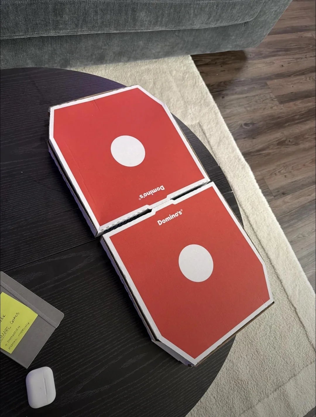 Two Domino's pizza boxes connected  a acheronian  circular  array  adjacent  to an AirPods lawsuit  and a grey tablet with a yellowish  sticky note