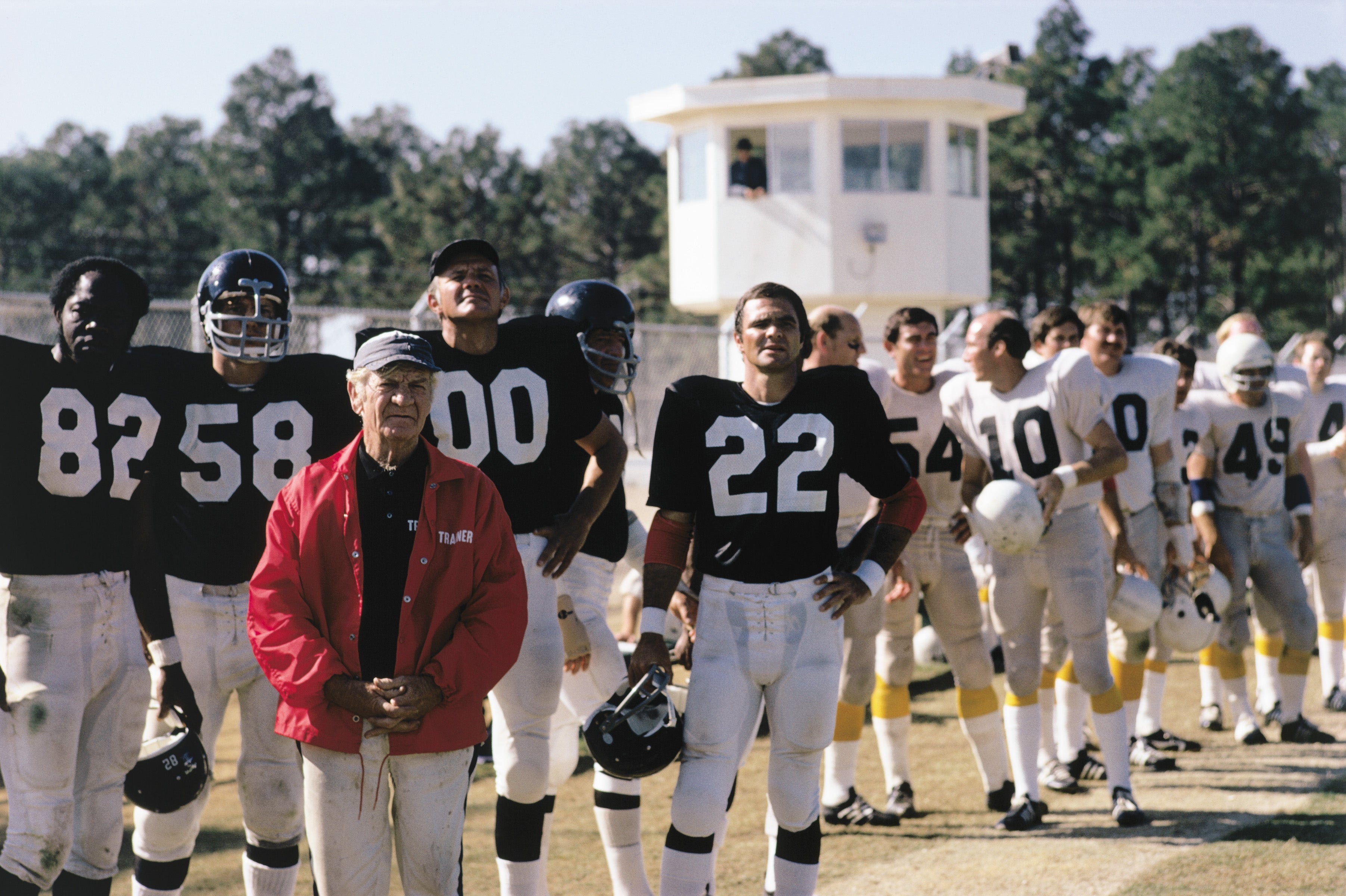 A football team stands on a field with a coach in a red jacket. Players in black and white jerseys are lined up, some holding helmets