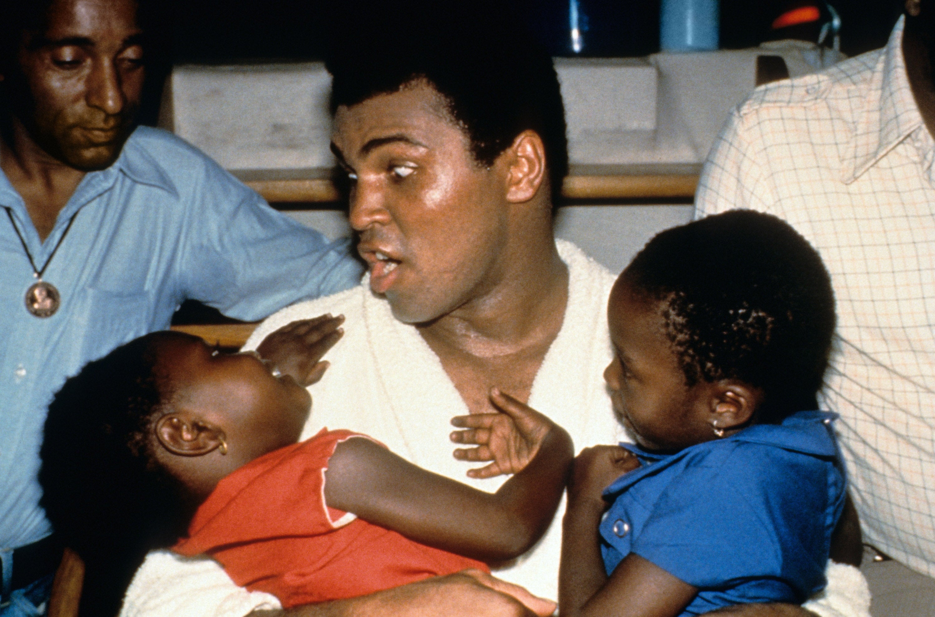 A renowned boxer, seated, holds two children in his arms, engaging with them warmly in a candid moment. People around him look on