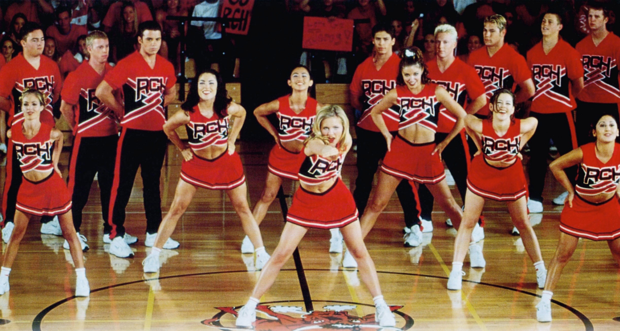 Cheerleading squad performs a routine on a gym floor. They're in coordinated outfits with bold "RCH" lettering on the front
