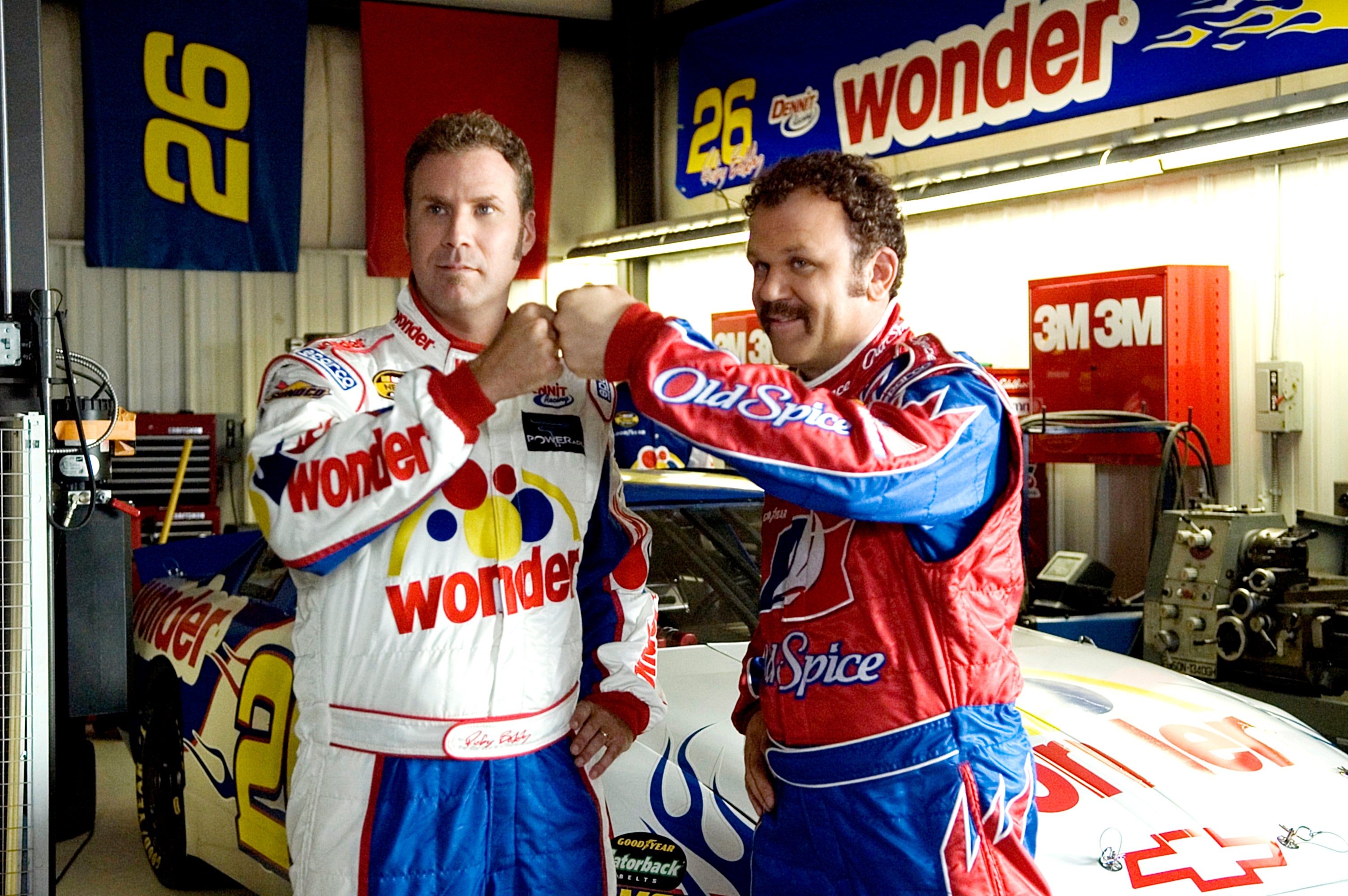 Two actors in racing suits fist bump in a garage with a race car in the background