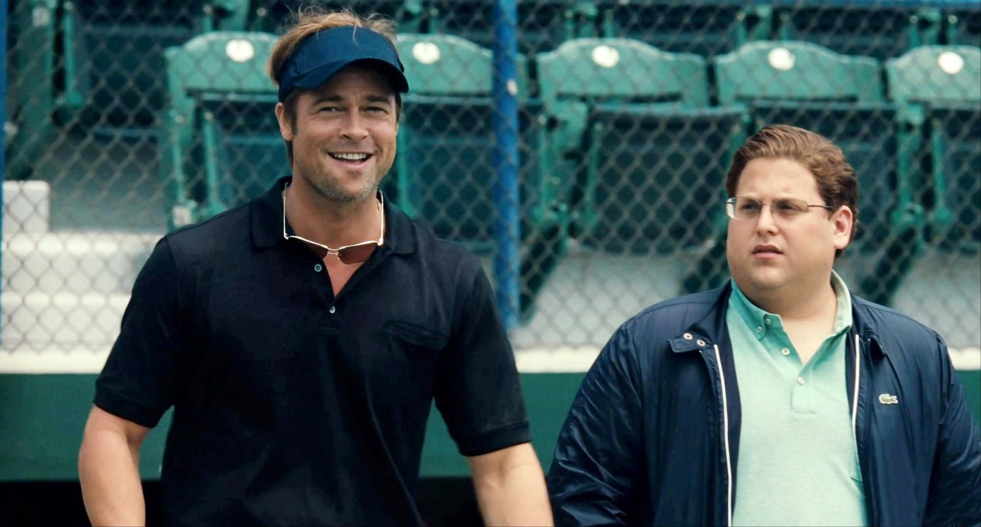 Two men from a scene in a sports setting, one wearing a visor and casual shirt, the other in a jacket and polo shirt, walking and talking