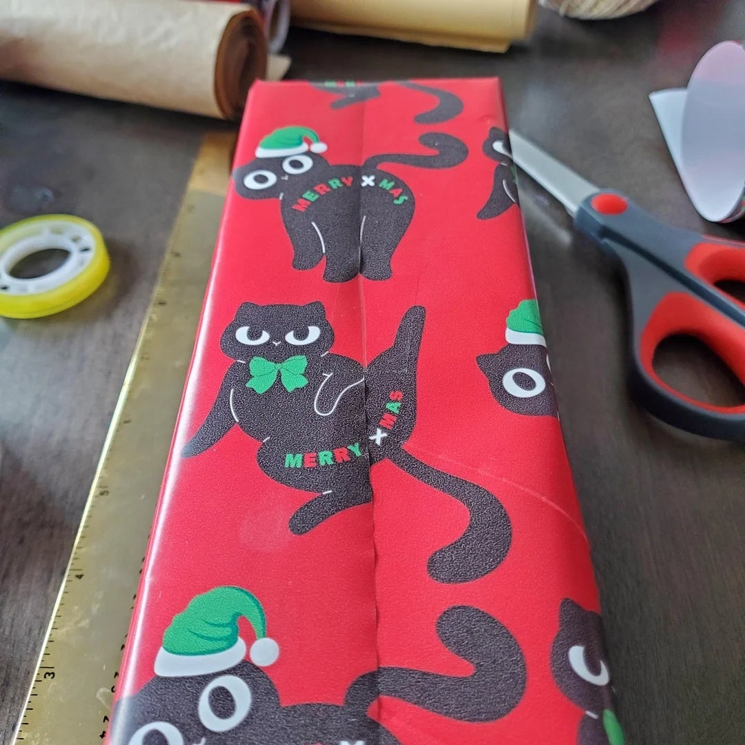 Gift wrapper  featuring cartoon achromatic  cats with Santa hats and "Merry Xmas" text, alongside wrapping tools similar  scissors and portion    connected  a table