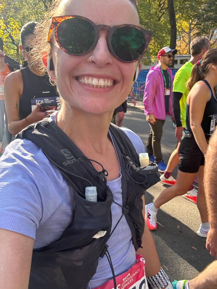 Person smiling during a marathon, wearing athletic gear and sunglasses, with runners in the background