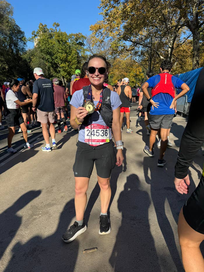 A person wearing athletic gear stands on a marathon course, holding a medal and smiling, surrounded by other participants