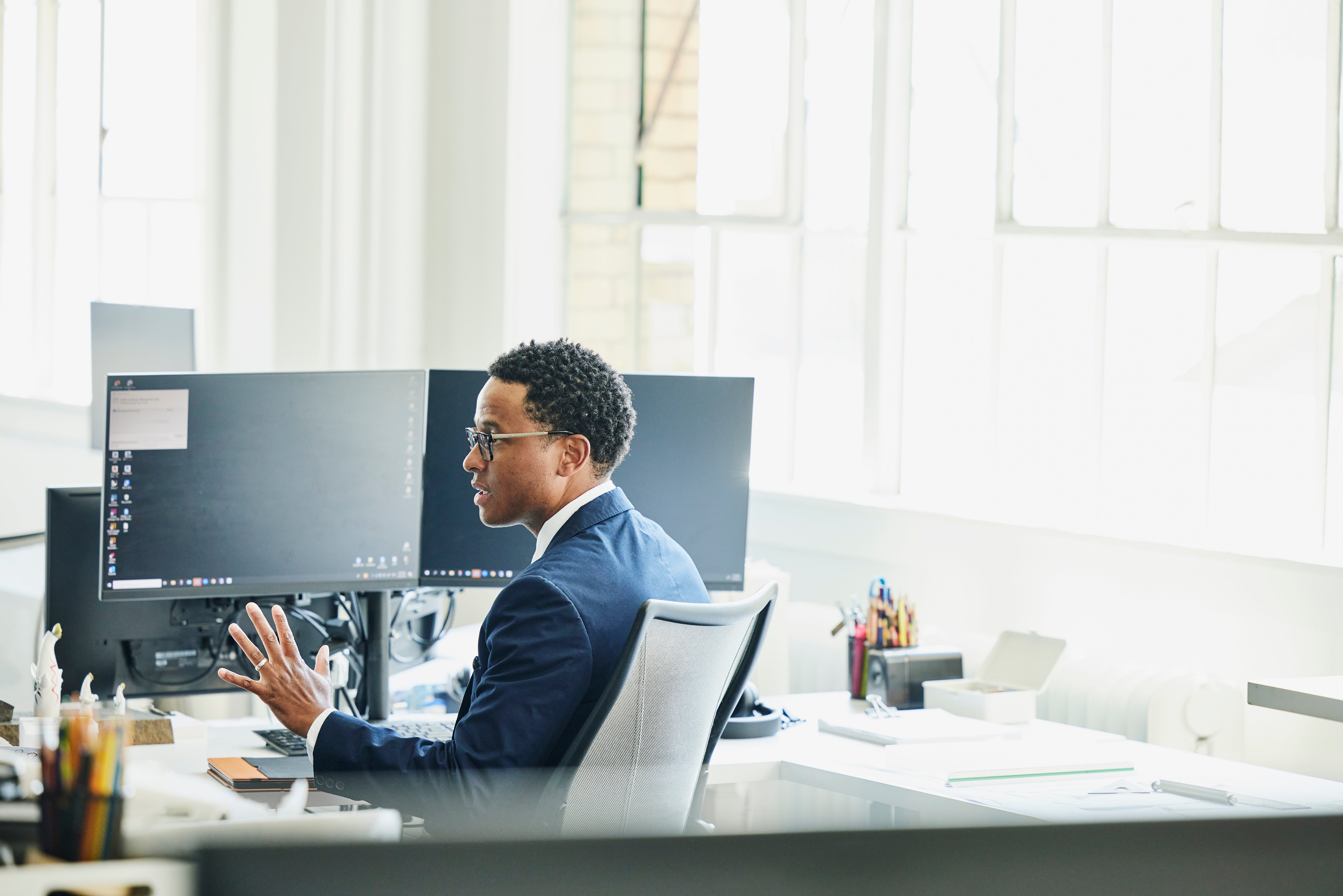 Person successful  a suit   sitting astatine  an bureau   desk, utilizing aggregate  machine  monitors. Bright country   with ample  windows